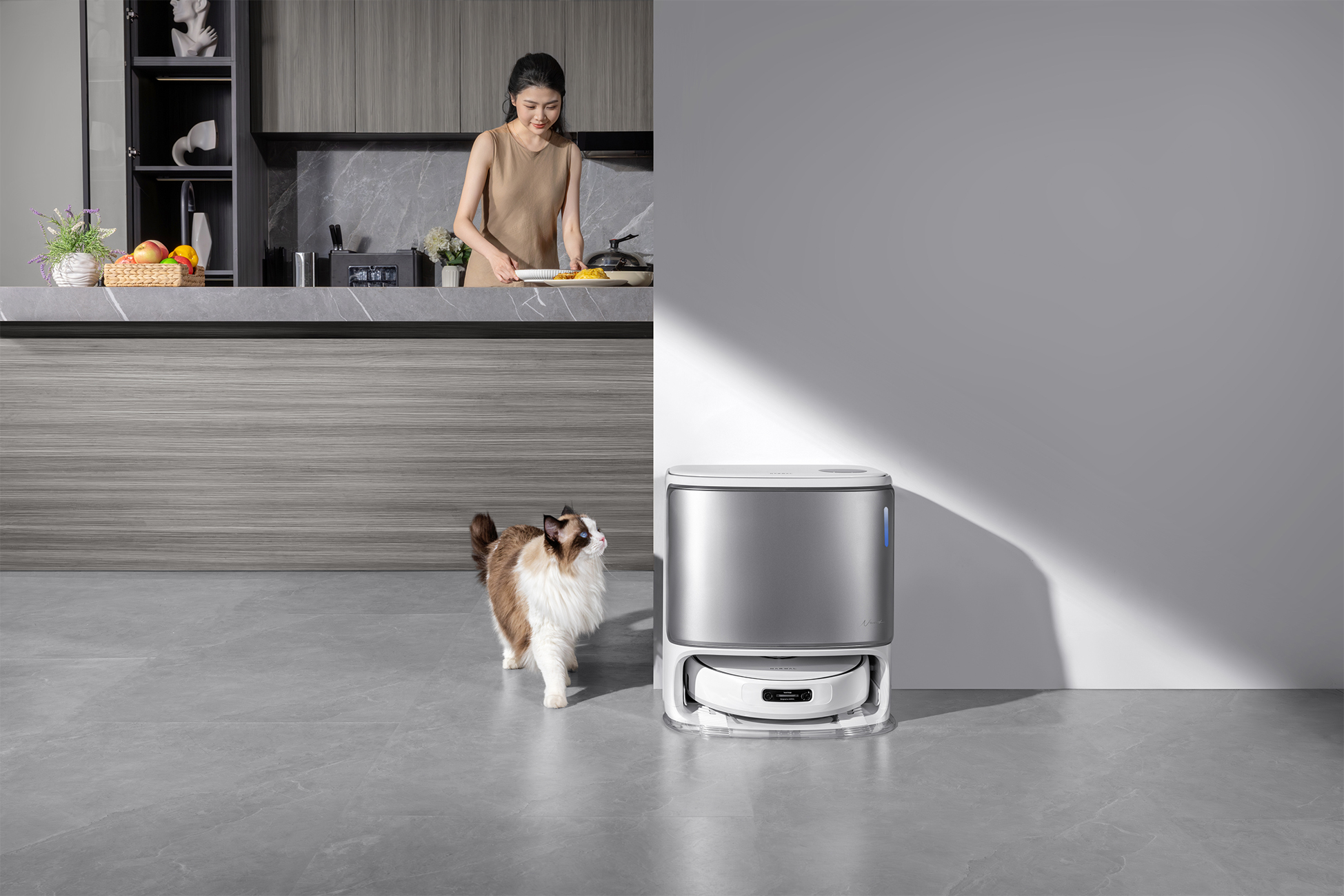Kobieta przygotowuje jedzenie w kuchni. Kot idzie w stronę robota sprzątającego. Scena jest jasna i nowoczesna.