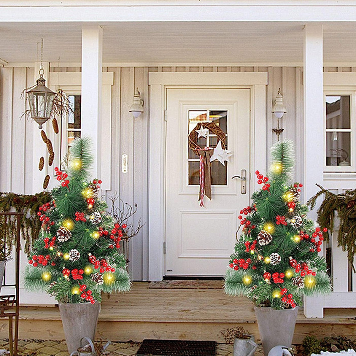 Zwei dekorierte Weihnachtsbäume in Töpfen flankieren eine weiße Tür mit einem Kranz. Die Veranda hat eine Laterne und festliche Dekoration.