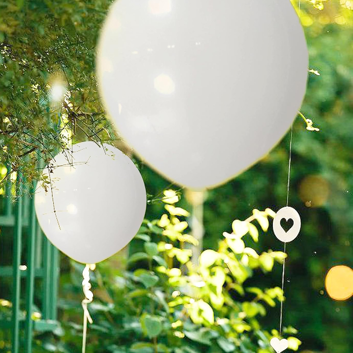 Zwei große weiße Ballons schweben inmitten grünen Laubes. Dekorative Herzen hängen an Schnüren, was auf eine Feier hindeutet.
