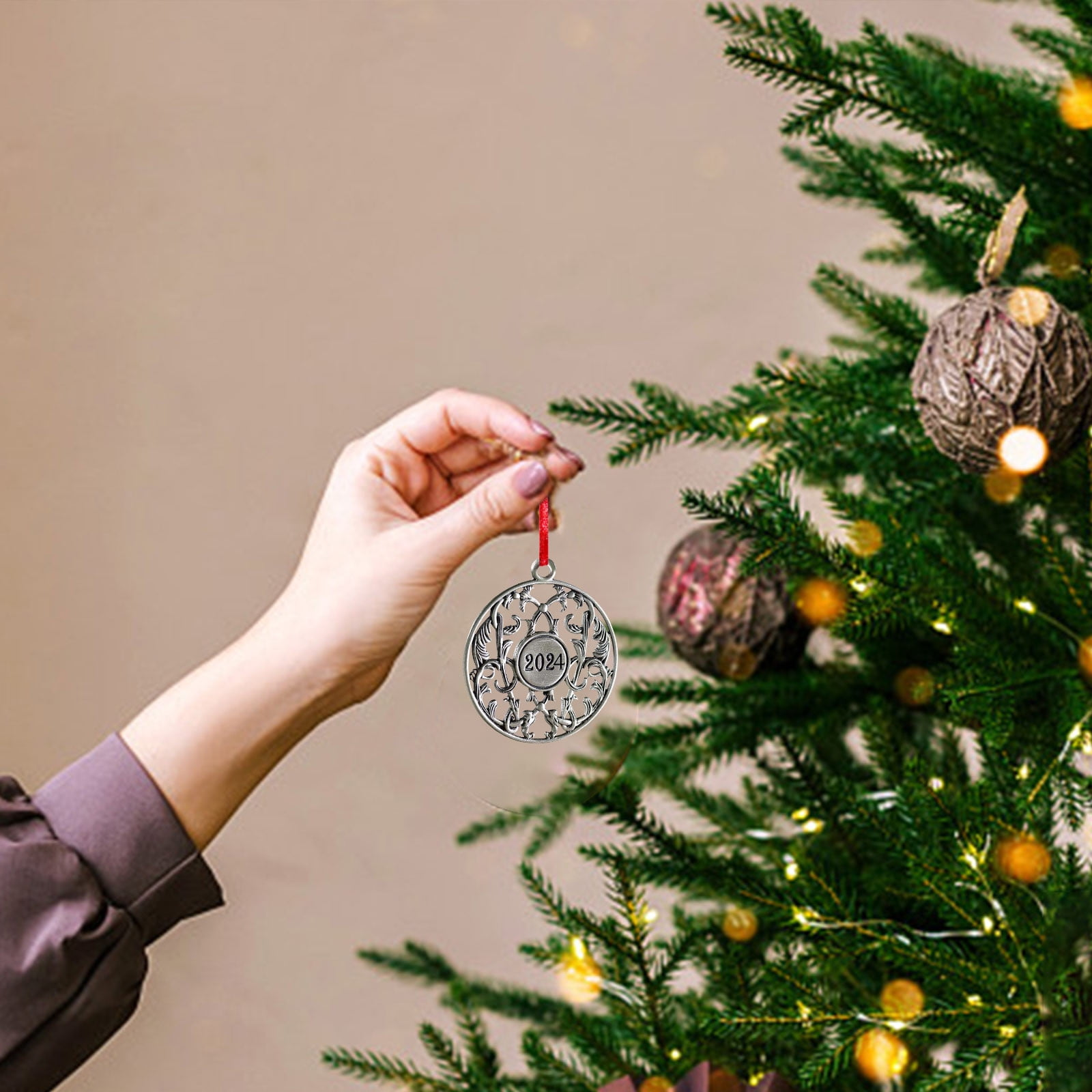 Eine Hand hält einen Weihnachtsbaumschmuck 2024. Ein Baum ist mit Ornamenten und Lichtern geschmückt. Beigefarbener Hintergrund.