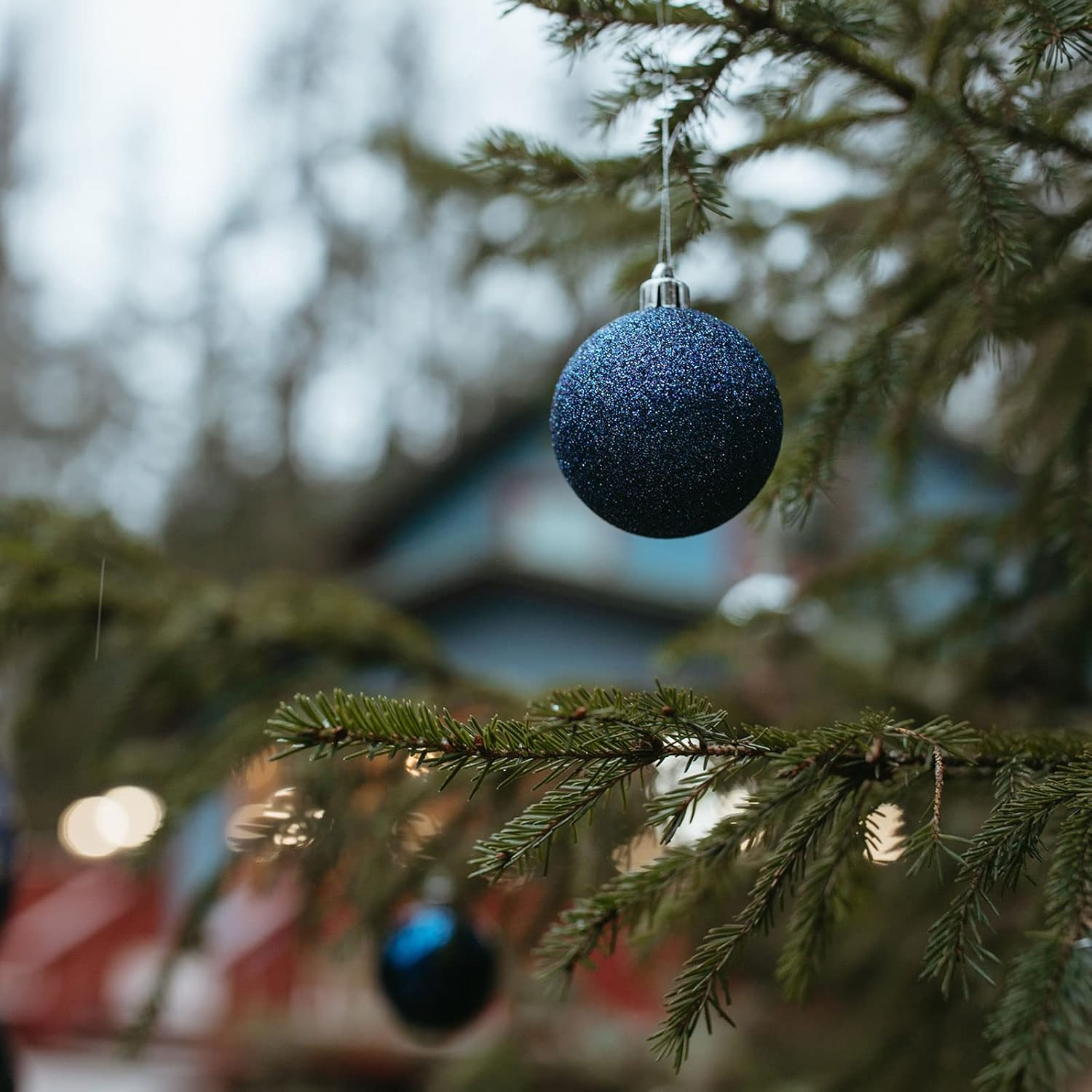 Nahaufnahme eines Weihnachtsbaumzweigs mit einer glitzernden blauen Kugel. Im Hintergrund ein verschwommenes Gebäude.