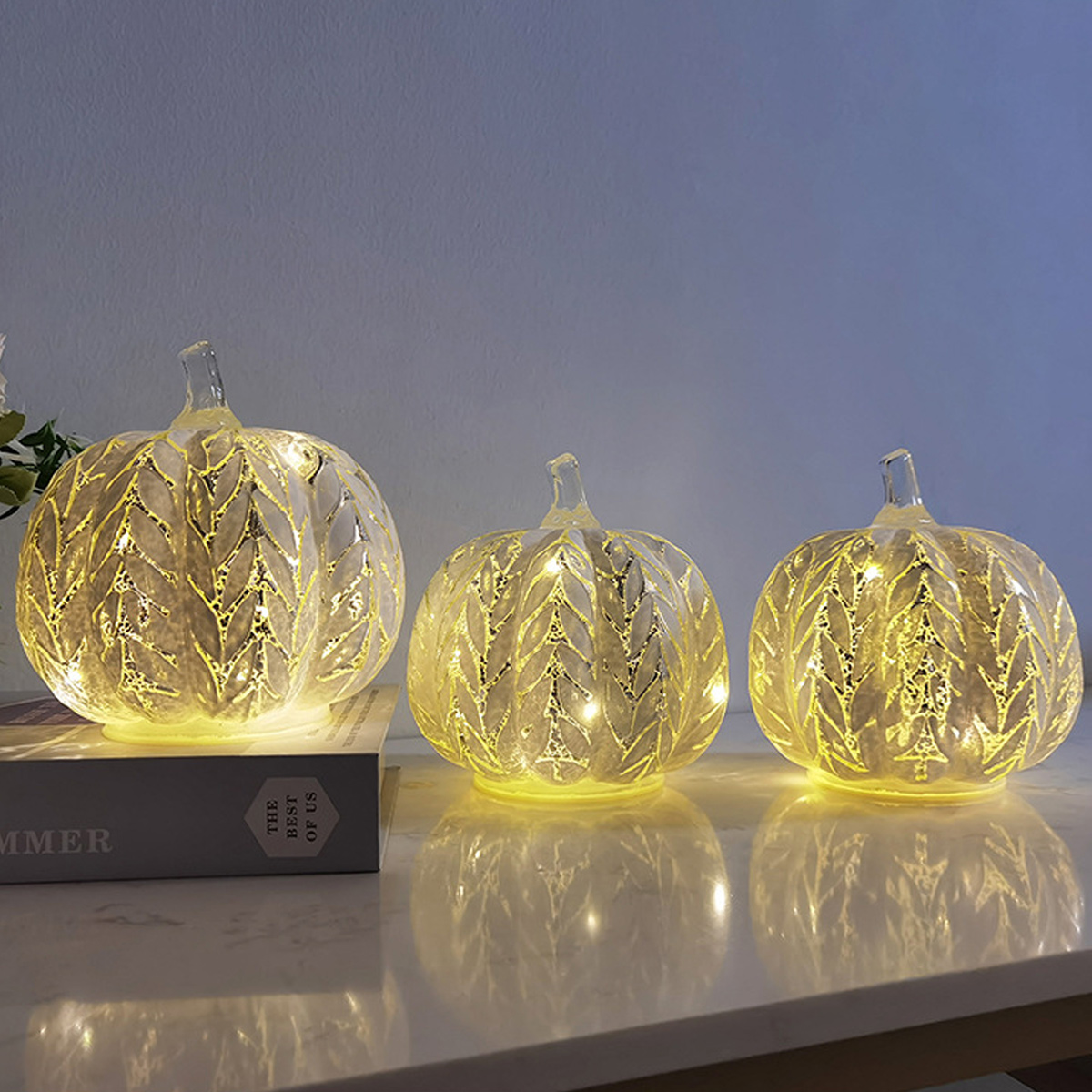 Drei beleuchtete Glaskürbisse mit Blättermustern stehen auf einem Tisch. Die Kürbisse leuchten warm und spiegeln sich.