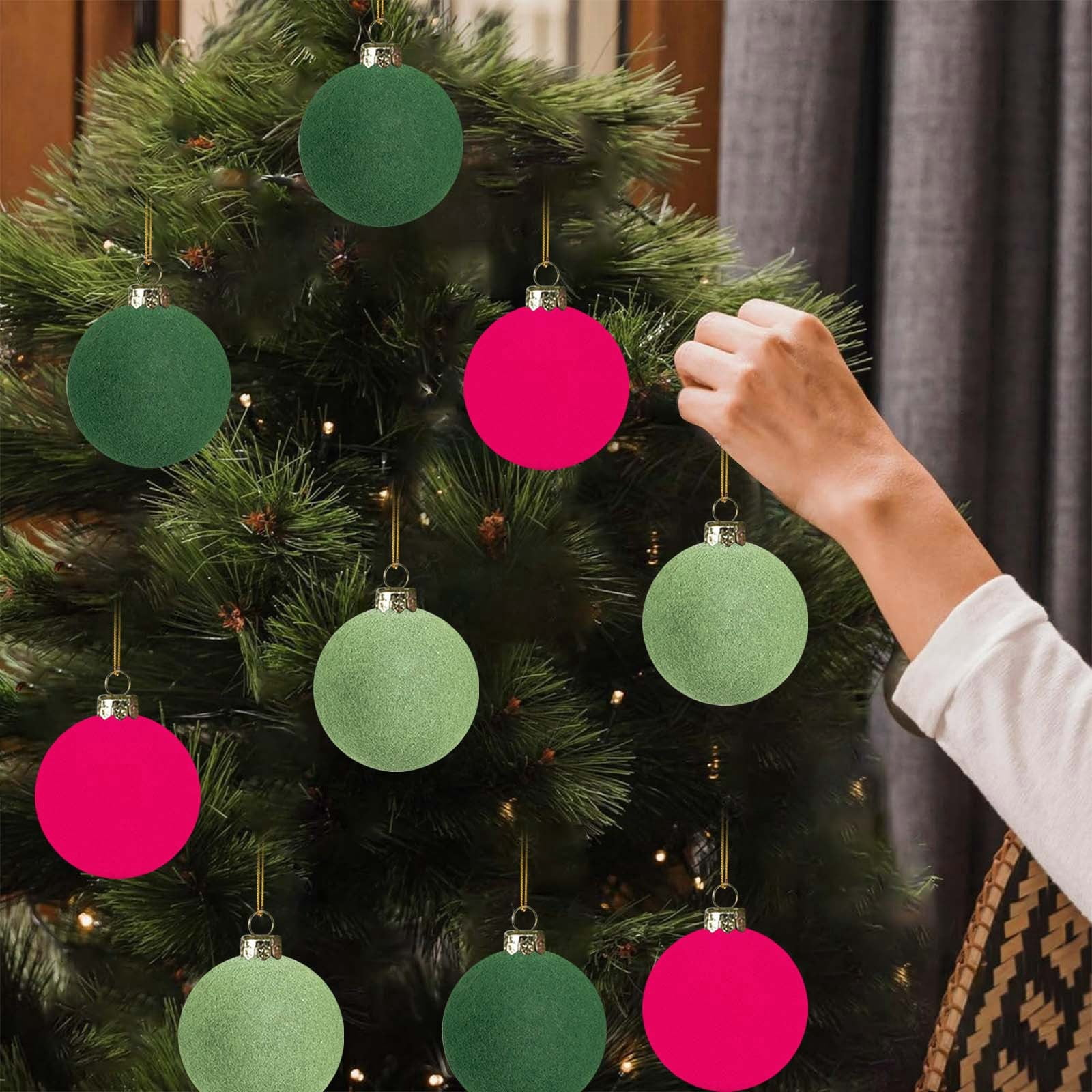 Eine Hand schmückt einen Weihnachtsbaum mit rosa und grünen Ornamenten. Der Hintergrund ist ein grauer Vorhang.