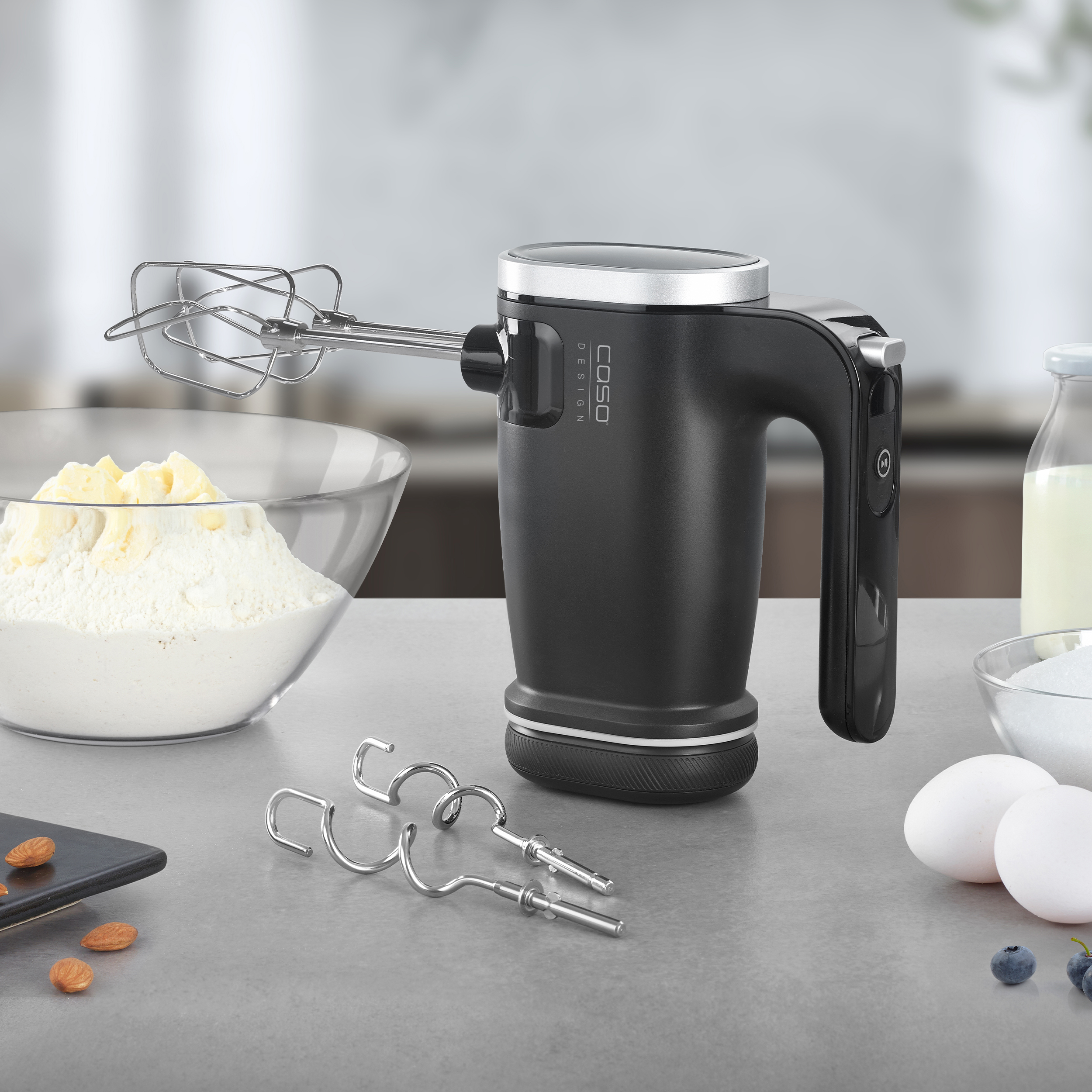 Un frullatore a mano nero con accessori, uova, farina e altri ingredienti per la cottura su un tavolo.