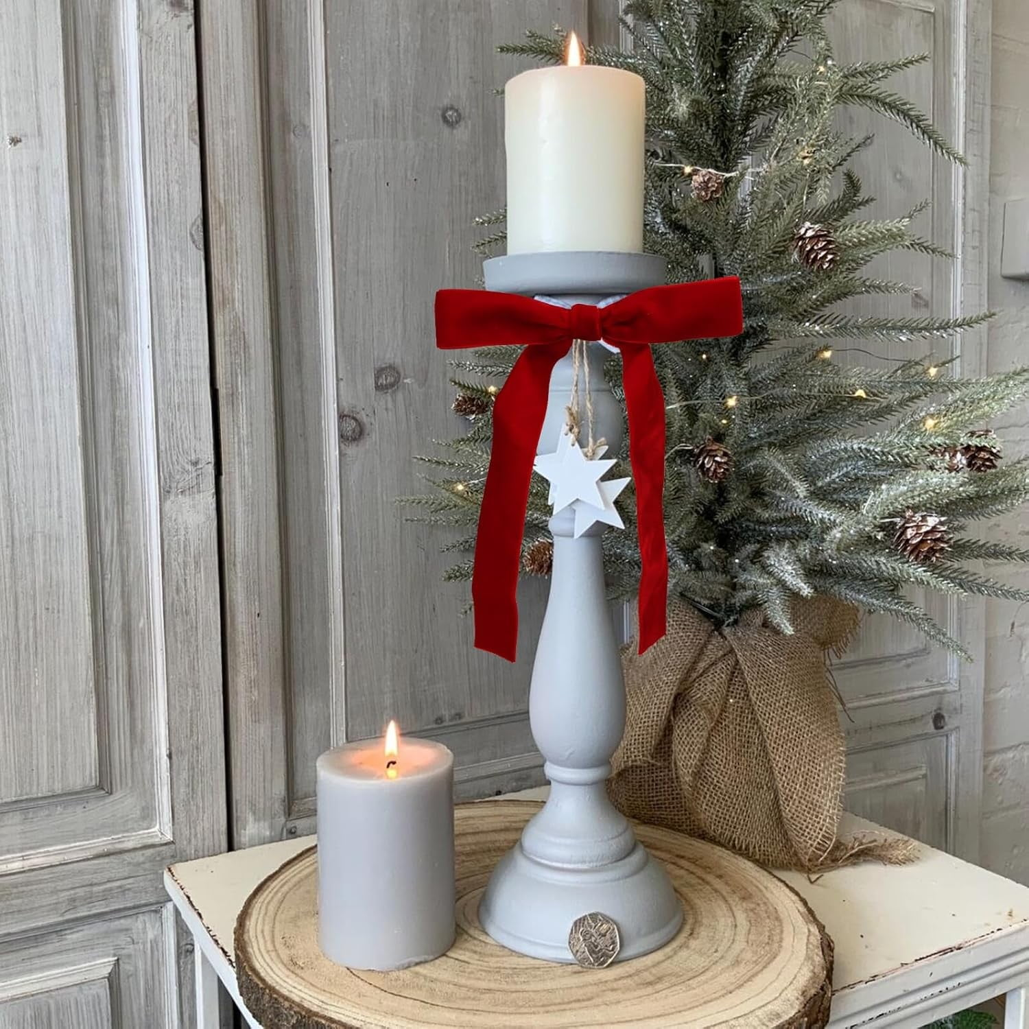 Eine brennende Kerze auf einem Ständer mit roter Schleife, eine kleinere Kerze und ein dekorierter Weihnachtsbaum im Hintergrund.