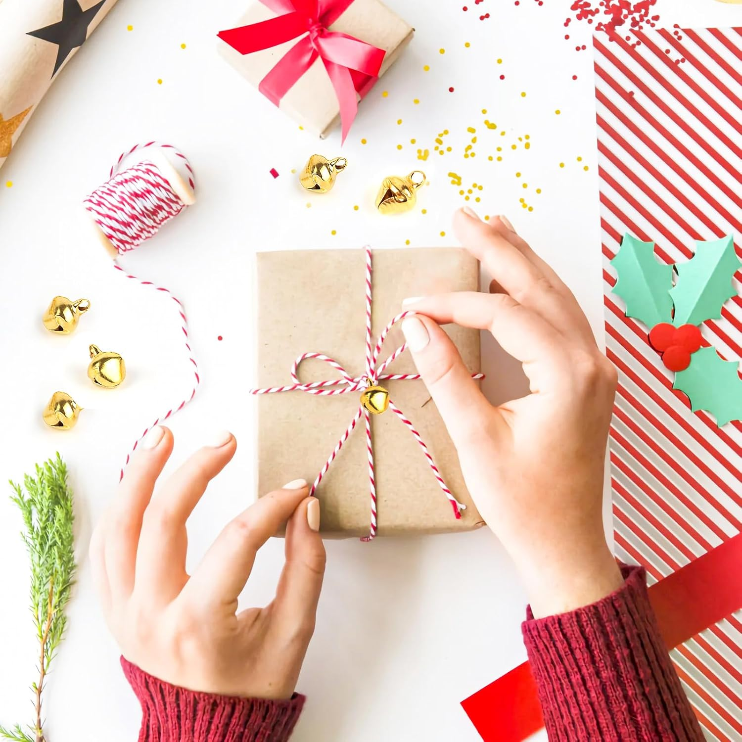 Hände beim Verpacken eines Geschenks mit Bindfaden und einer kleinen Glocke. Andere Geschenke und Weihnachtsdekorationen sind sichtbar.