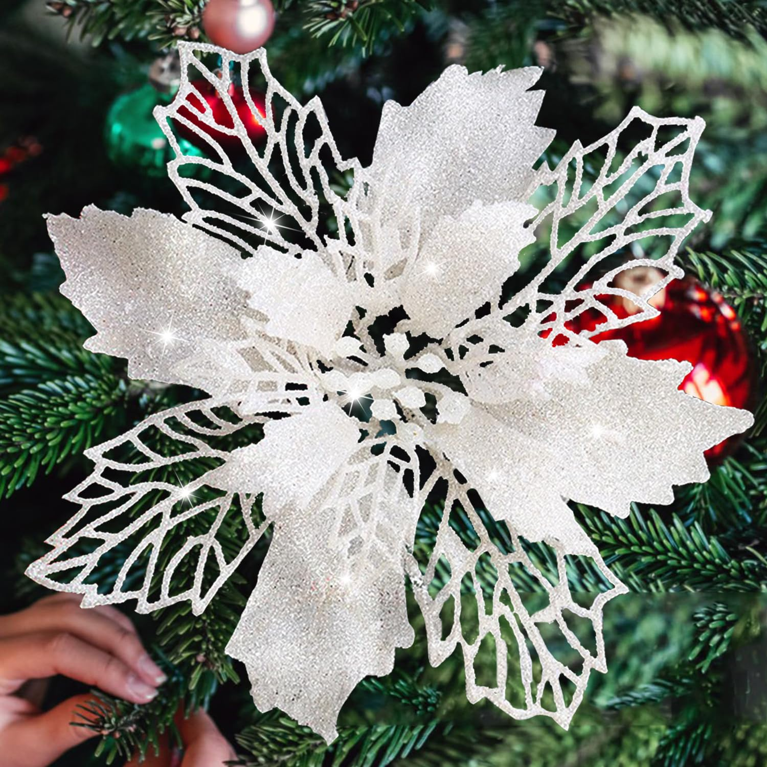 Ein glitzernder, weißer Weihnachtsstern auf einem Weihnachtsbaum. Rote, grüne und rosa Ornamente im Hintergrund. Eine Hand justiert die Blume.