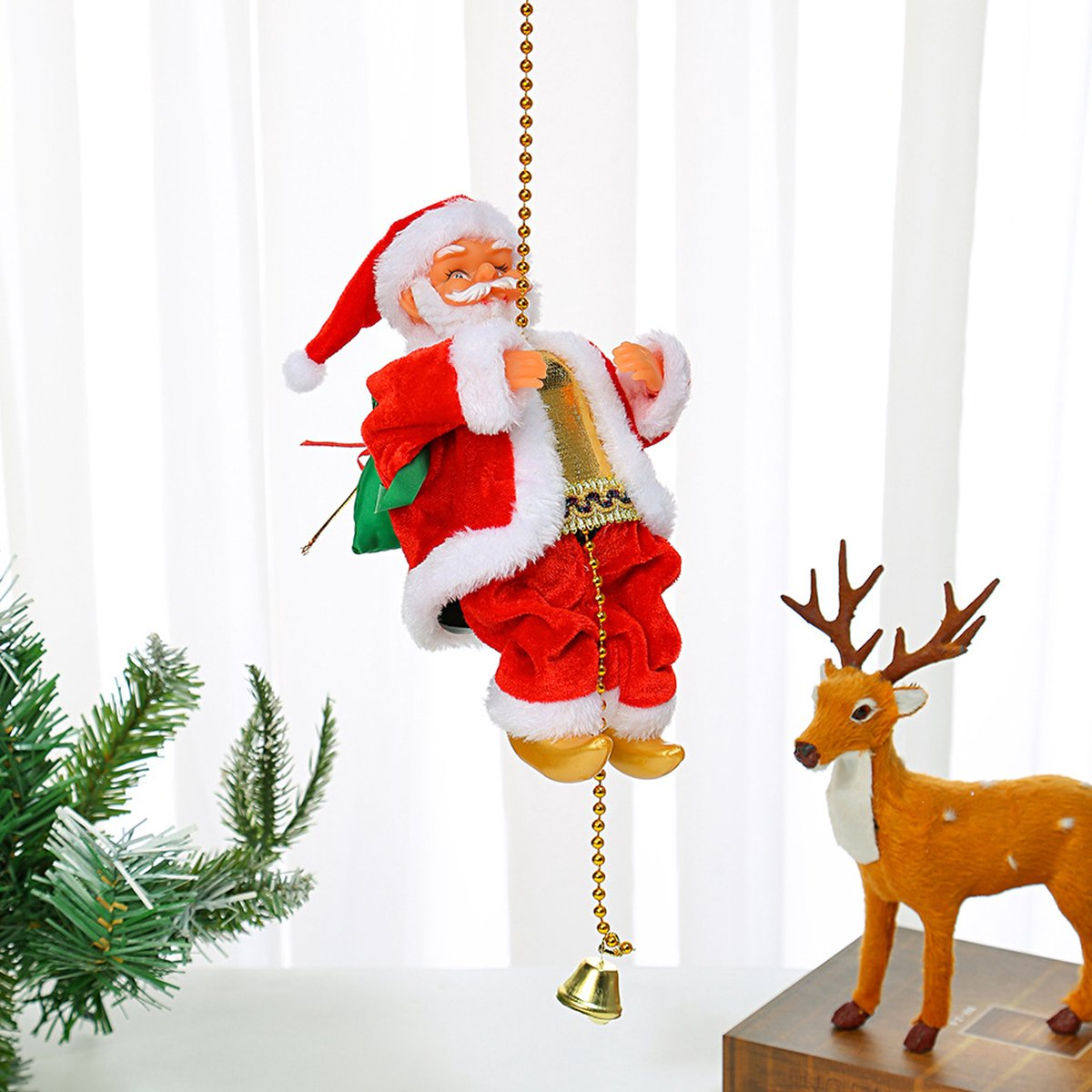 Eine Weihnachtsmann-Puppe hängt an einer Kette. Rechts steht eine Rentierfigur. Im unteren linken Bereich befinden sich grüne Zweige.