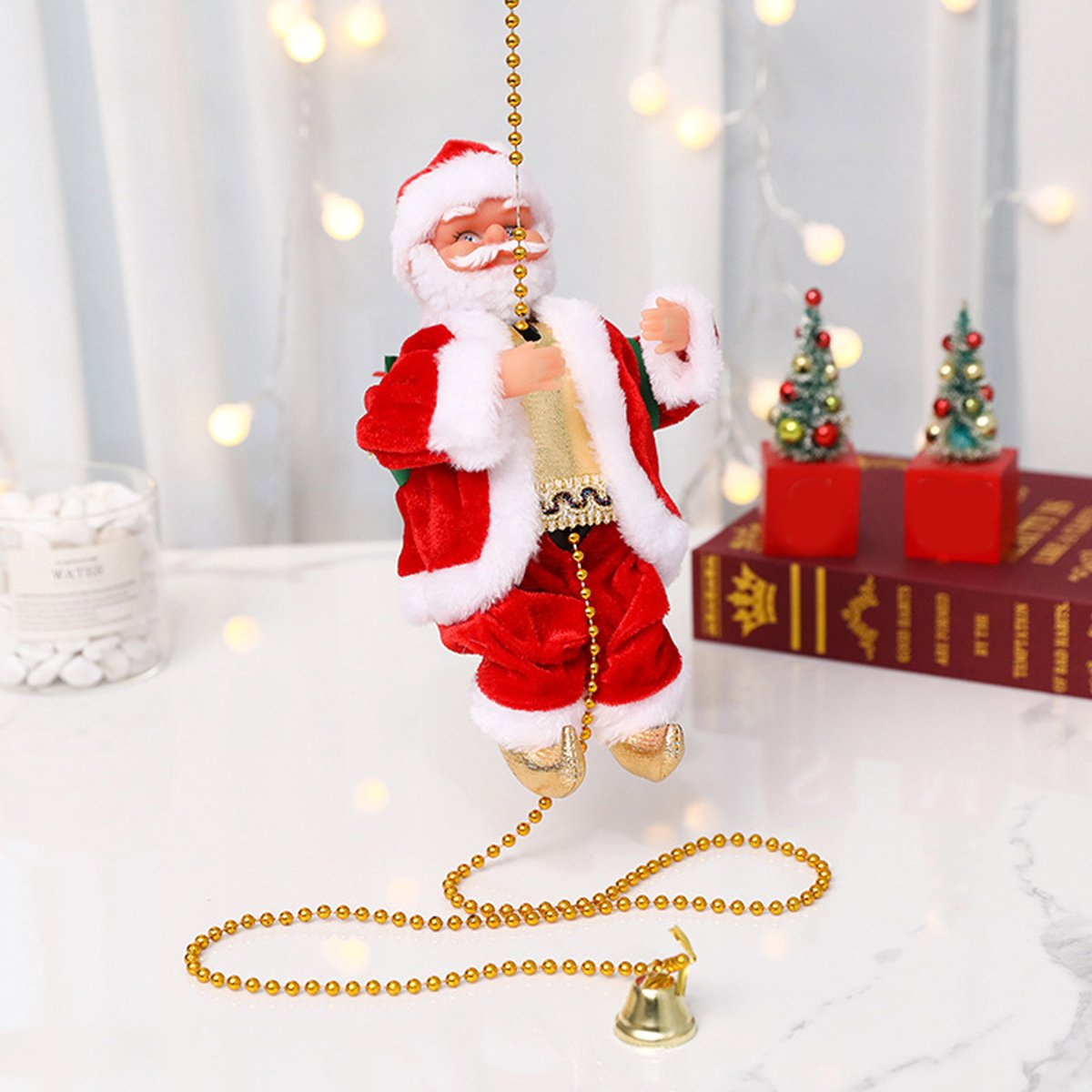 Eine Weihnachtsmannpuppe hängt an einer Goldkette, im Hintergrund kleine Weihnachtsbäume und Bücher. Am Ende der Kette hängt eine Glocke.