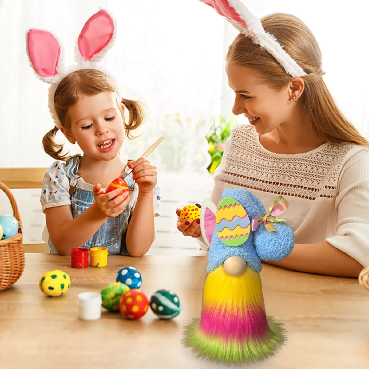 Ein Mädchen und eine Frau bemalen Ostereier an einem Tisch, beide tragen Hasenohren. Osterzwerg ist vorne. Bunte Eier und Farbe.