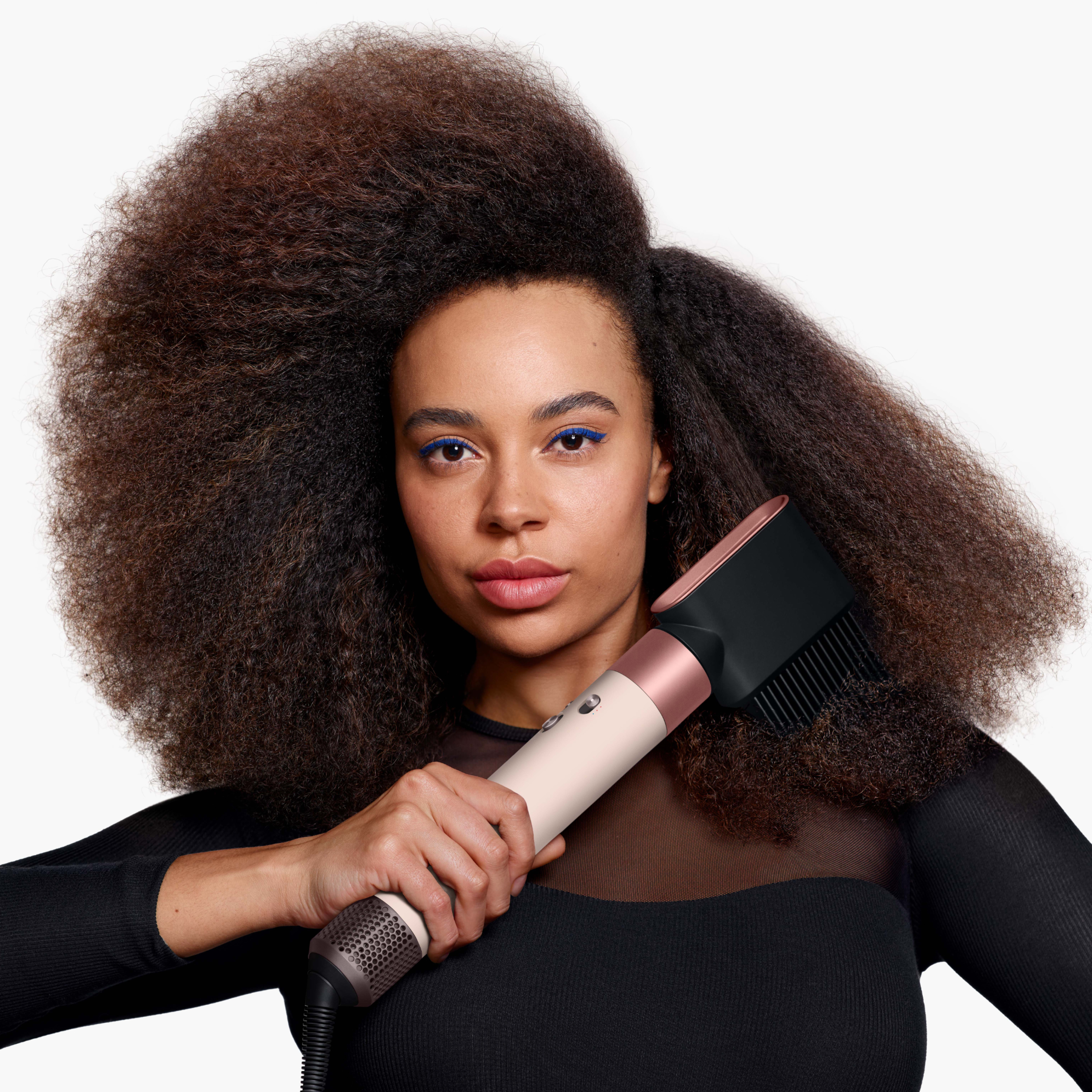 Eine Frau hält einen Haartrockner. Sie hat dunkle Haut, blauen Eyeliner und großes, lockiges Haar. Sie trägt ein schwarzes Oberteil.