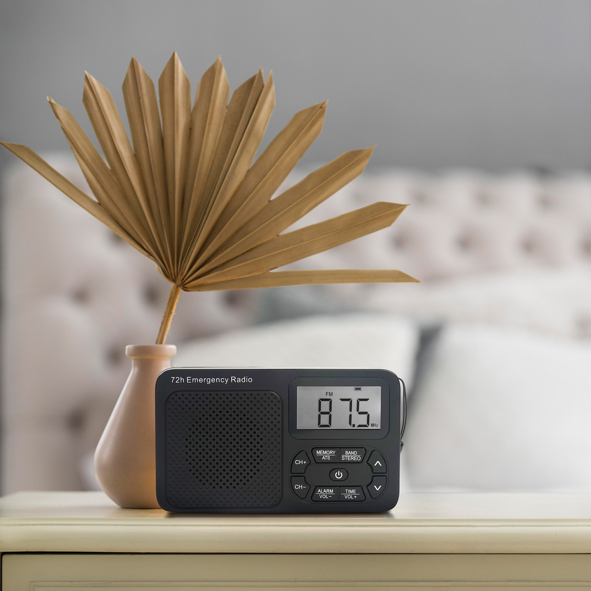 A black emergency radio on a table. A vase with dried leaves is behind it. A bed is in the background.
