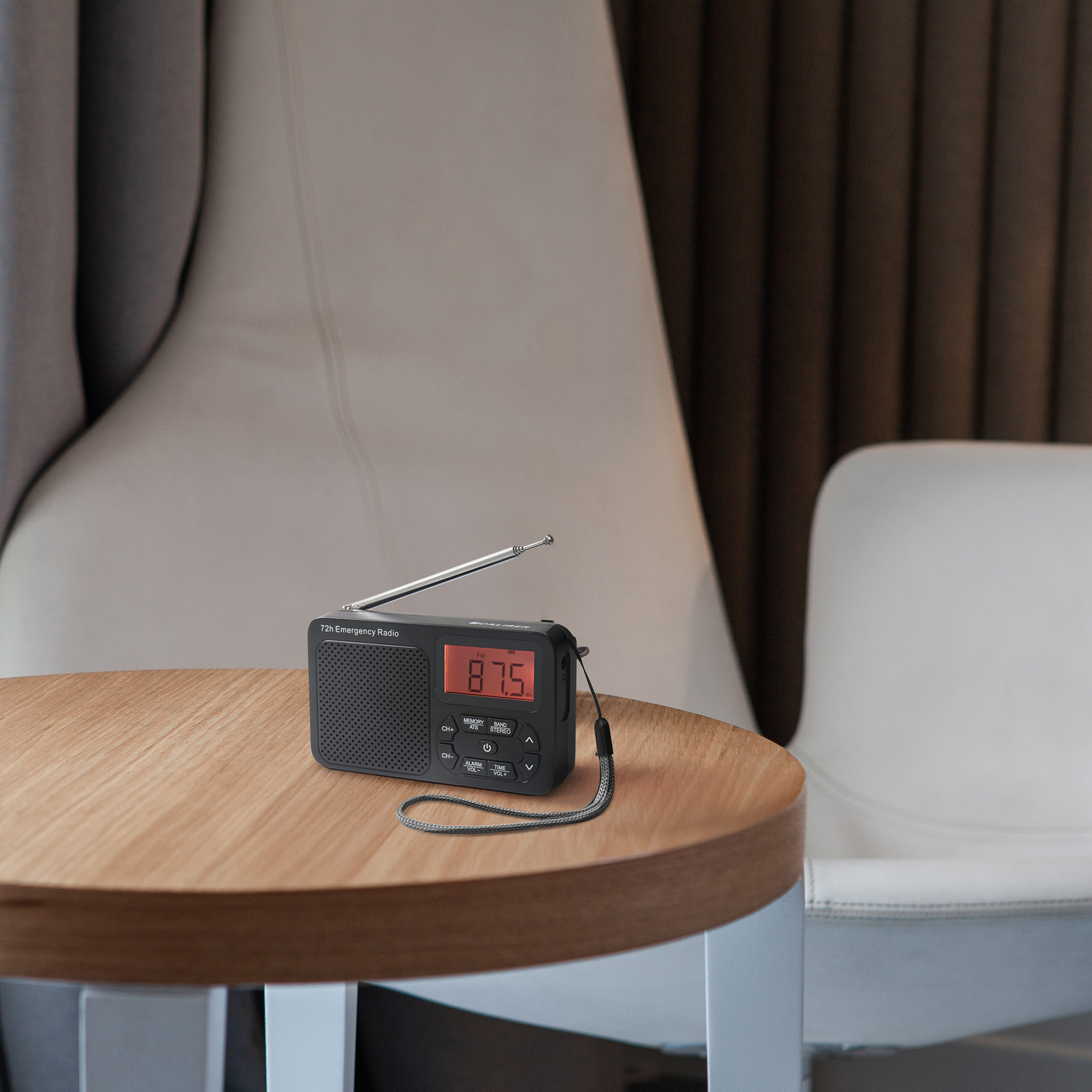 A black emergency radio on a wooden table. The radio displays 87.5 MHz. Chairs and curtains are in the background.