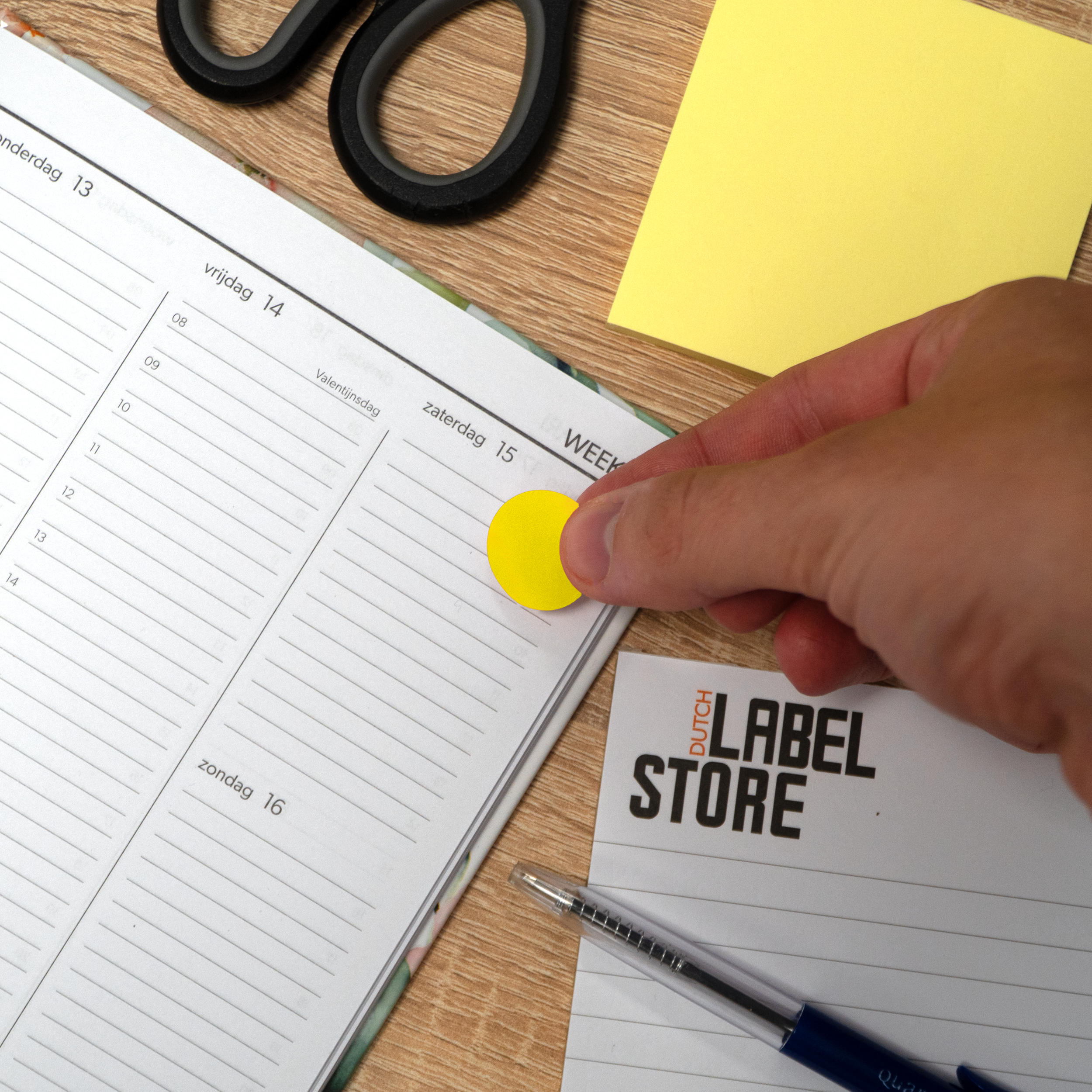 Een hand plaatst een gele plakbriefje op een planner. Schaar en andere notities zijn ook aanwezig.