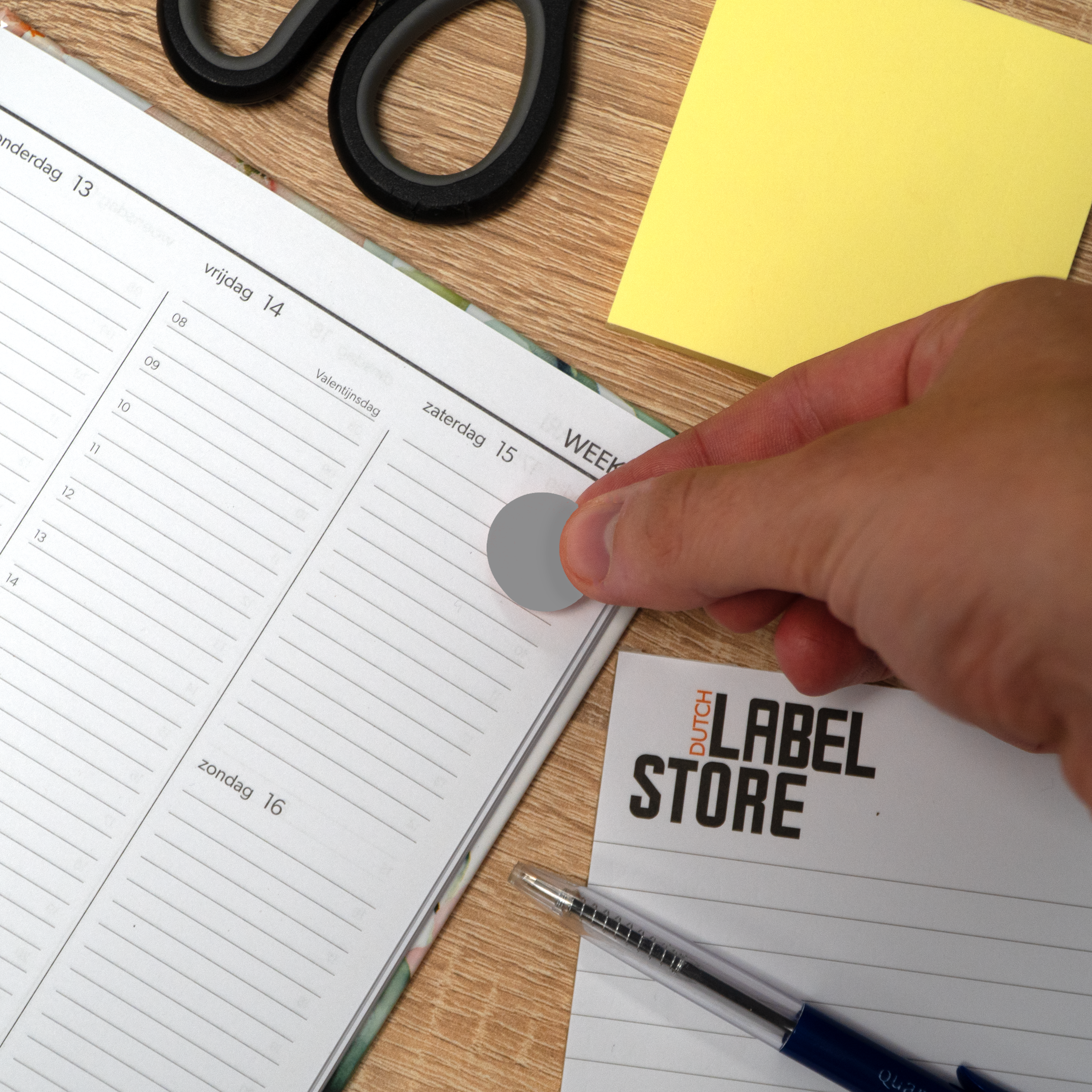 Een hand plaatst een grijze sticker op een open planner. Schaar, gele notitie en pen zijn ook zichtbaar.
