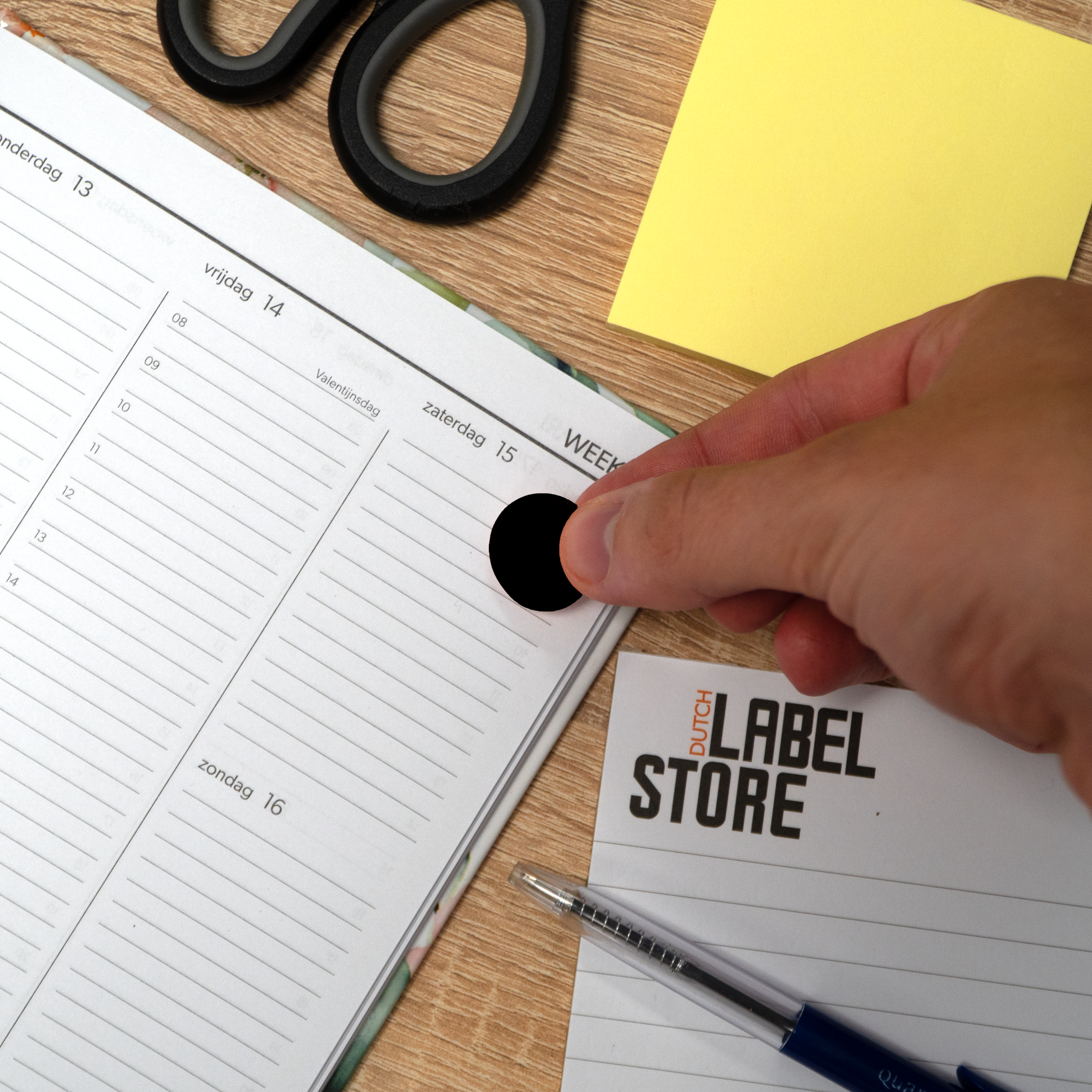 Een hand plaatst een zwarte sticker op een planner. Schaar en een gele plakbriefje liggen op een houten tafel.