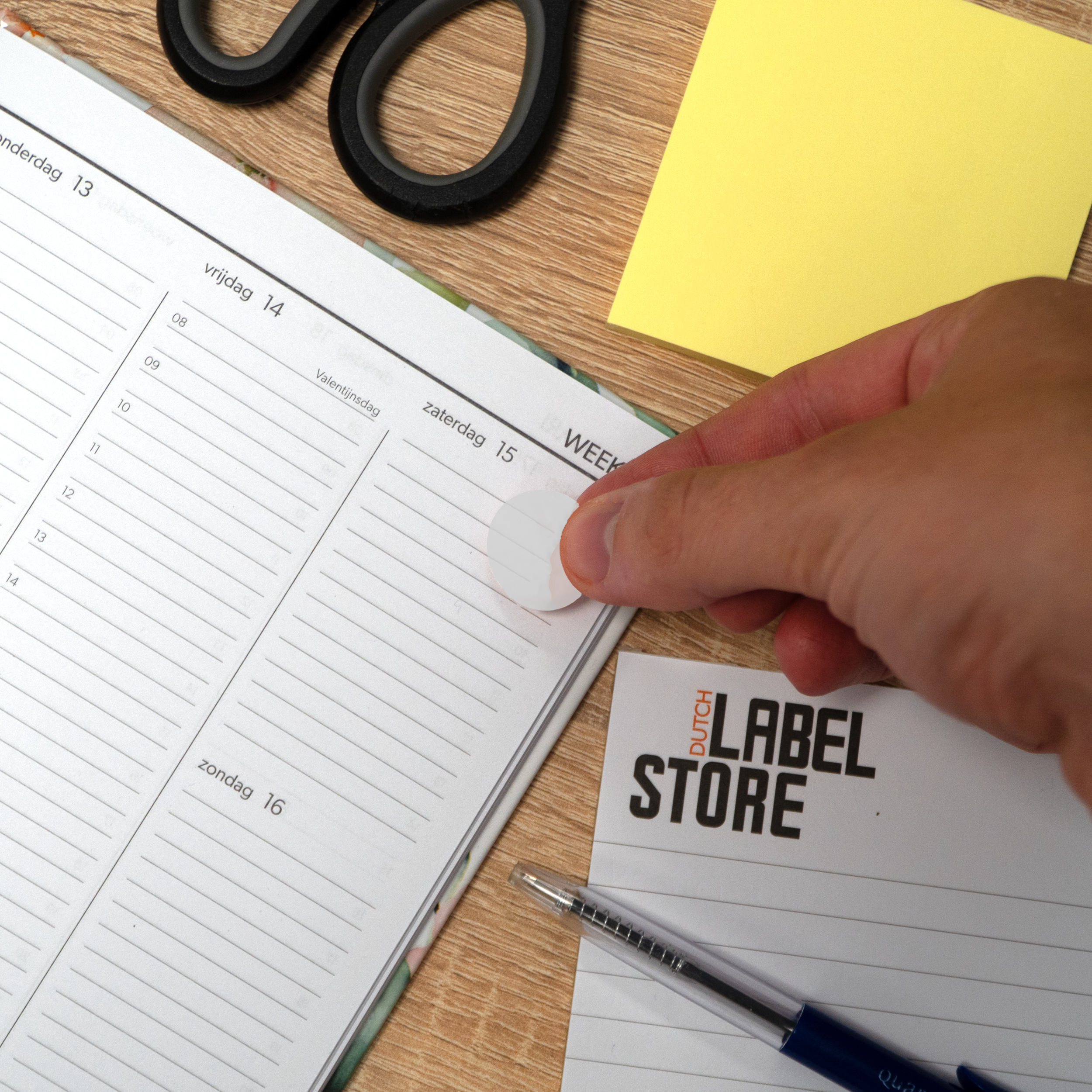 Een hand plakt een sticker op een planner. Schaar en gele notitie op houten oppervlak.