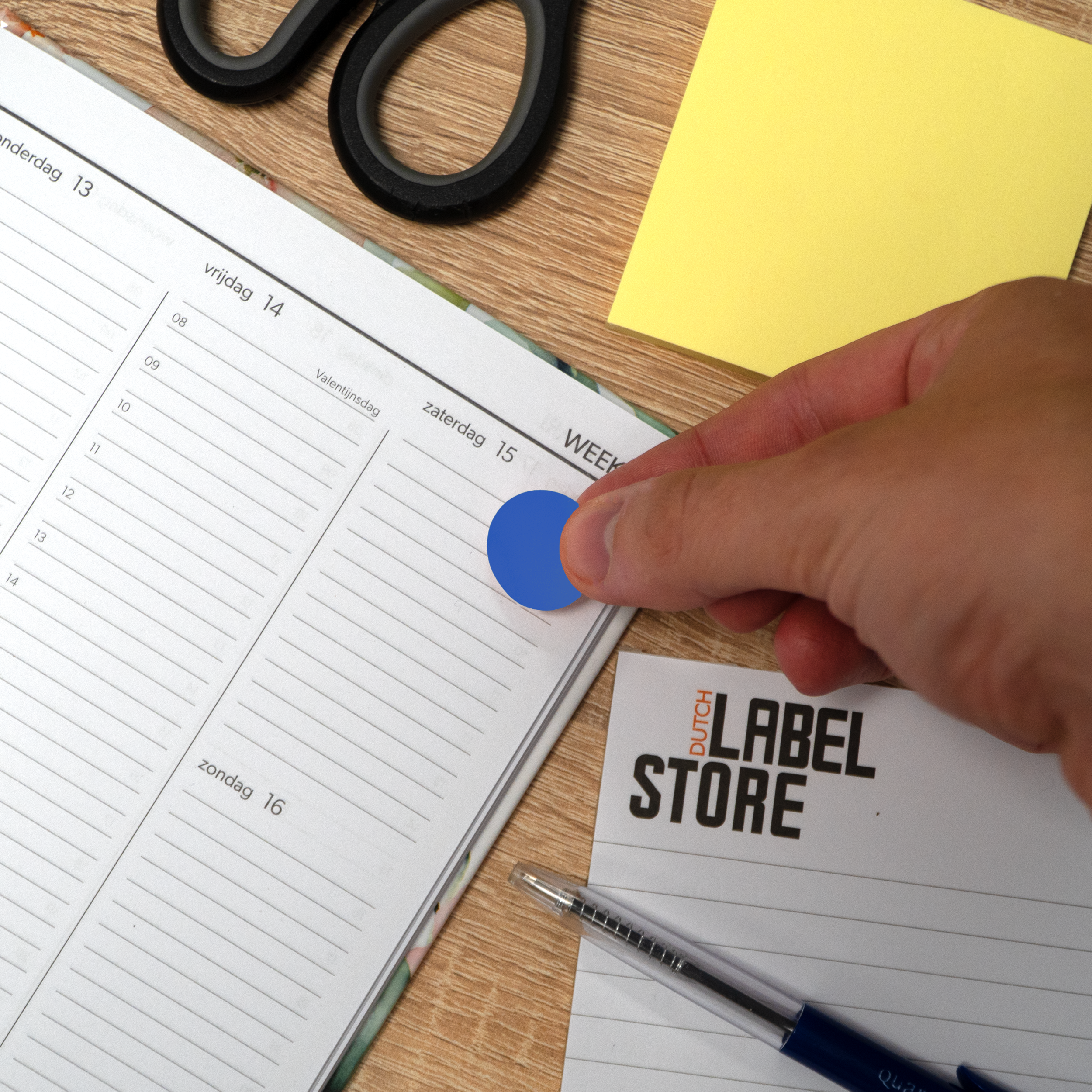 Een hand plaatst een blauwe sticker op een planner. Schaar, gele plakbriefje en Dutch Label Store zijn zichtbaar.