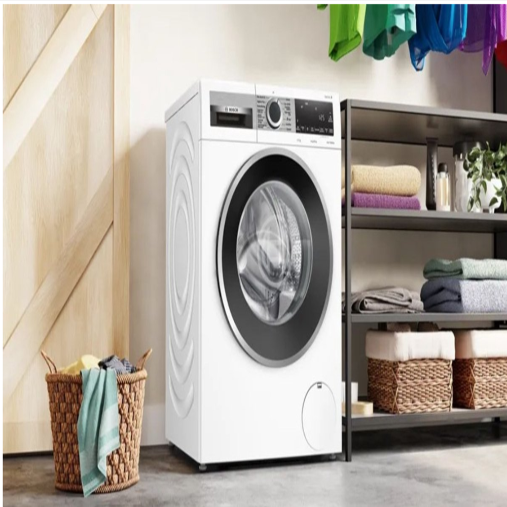 White washing machine with a round black door, next to a wooden door and a basket of clothes. Shelves with towels are in the background.