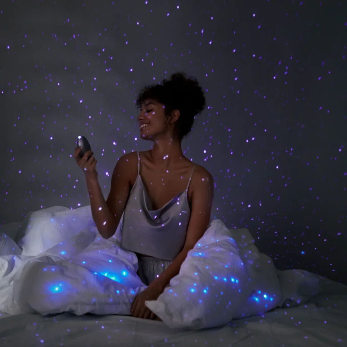 Kobieta w piżamie na łóżku, patrzy na projektor gwiazd. Fioletowe gwiazdy rzucane na nią i pościel. Ciemne tło.