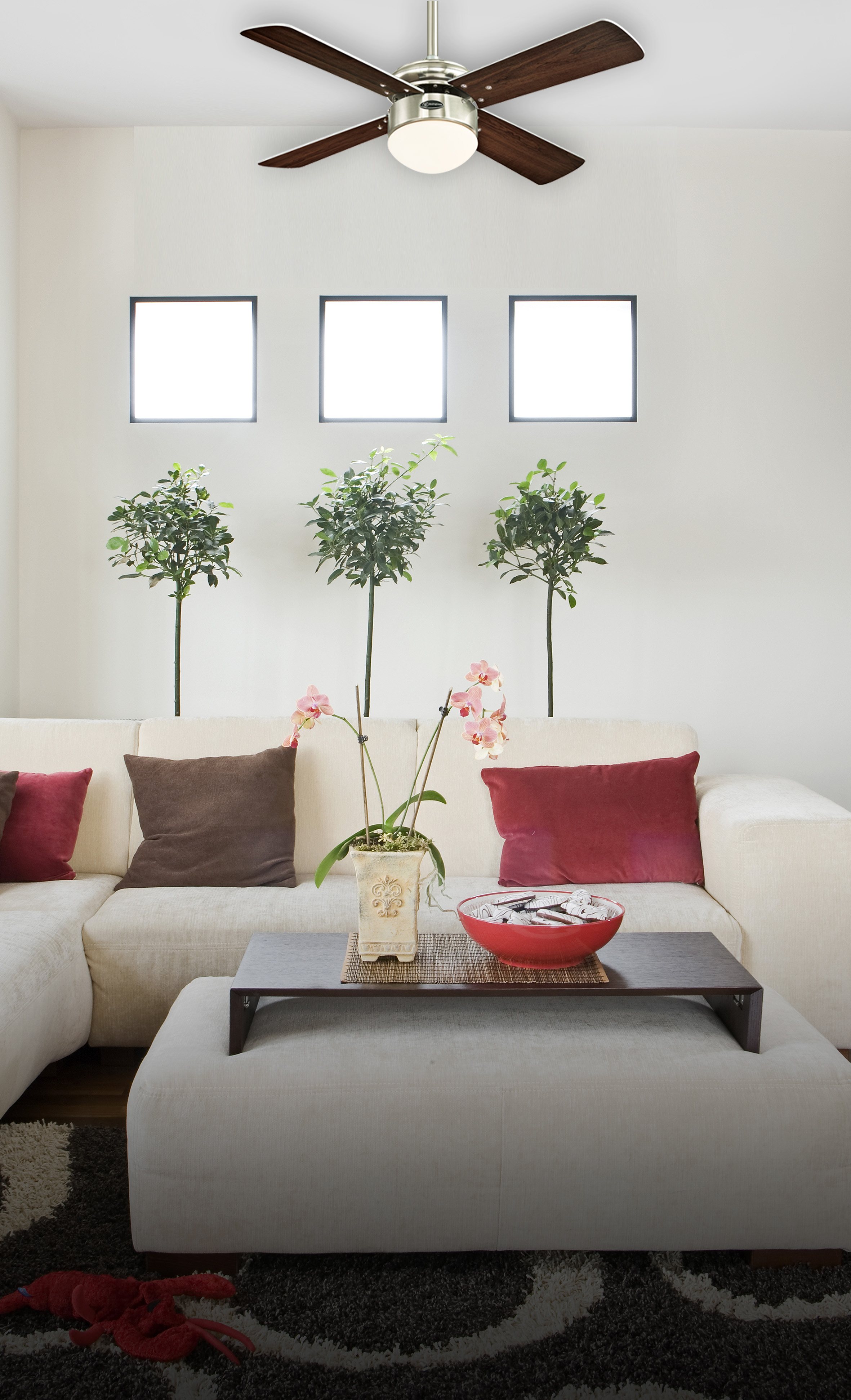 Wohnzimmer mit Sofa, Pflanzen und Deckenventilator. Ein niedriger Tisch trägt eine Schale. Fenster und ein Teppich sind sichtbar.