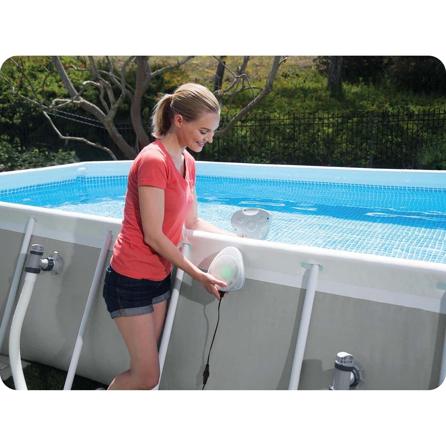 Una mujer con pantalones cortos y una camiseta roja ajusta un limpiador de piscina en una piscina rectangular. Hay árboles y una valla en el fondo.