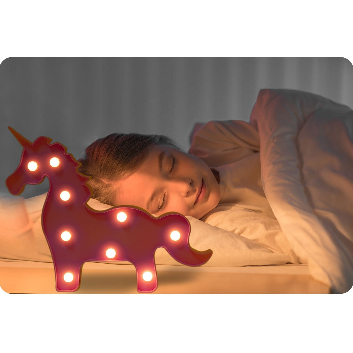 Una niña duerme junto a una luz con forma de unicornio. La luz es rosa con bombillas blancas. El fondo es una cortina gris suave.