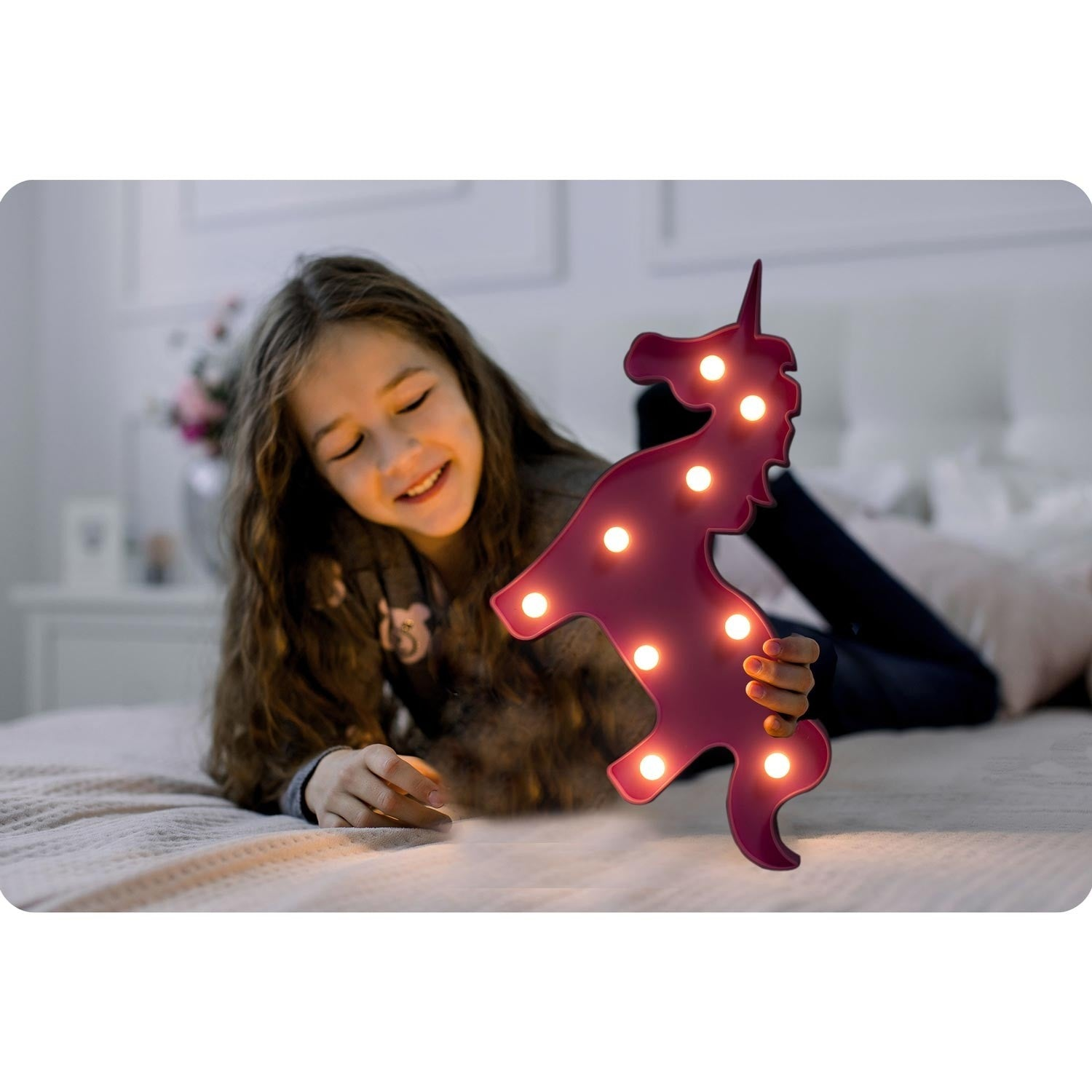 Una niña sonríe, sosteniendo una luz de unicornio rosa en una cama. El unicornio tiene luces brillantes. El fondo muestra una habitación y flores.