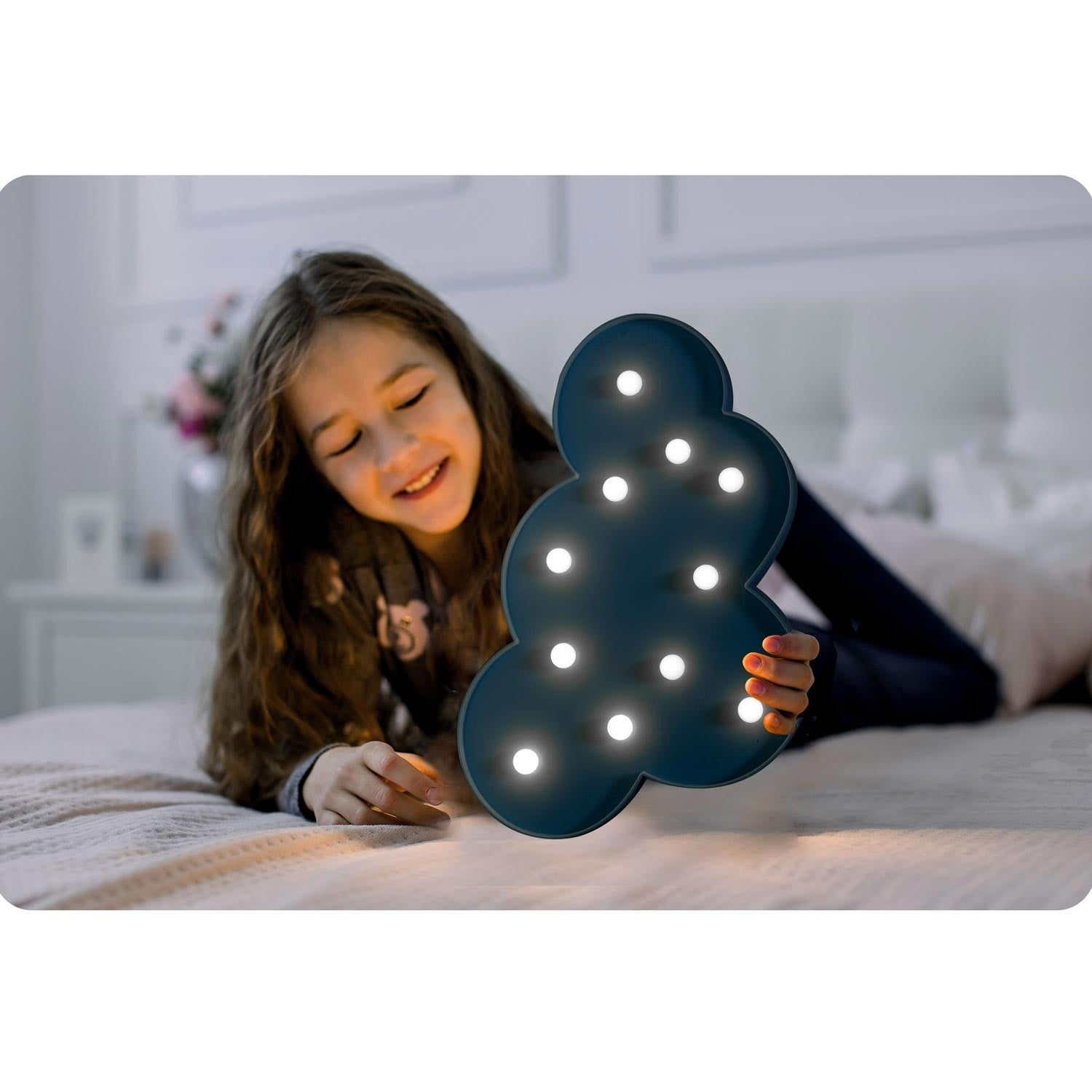 Una niña sonríe, sosteniendo una luz en forma de nube. Está acostada en una cama. La luz emite un brillo suave.