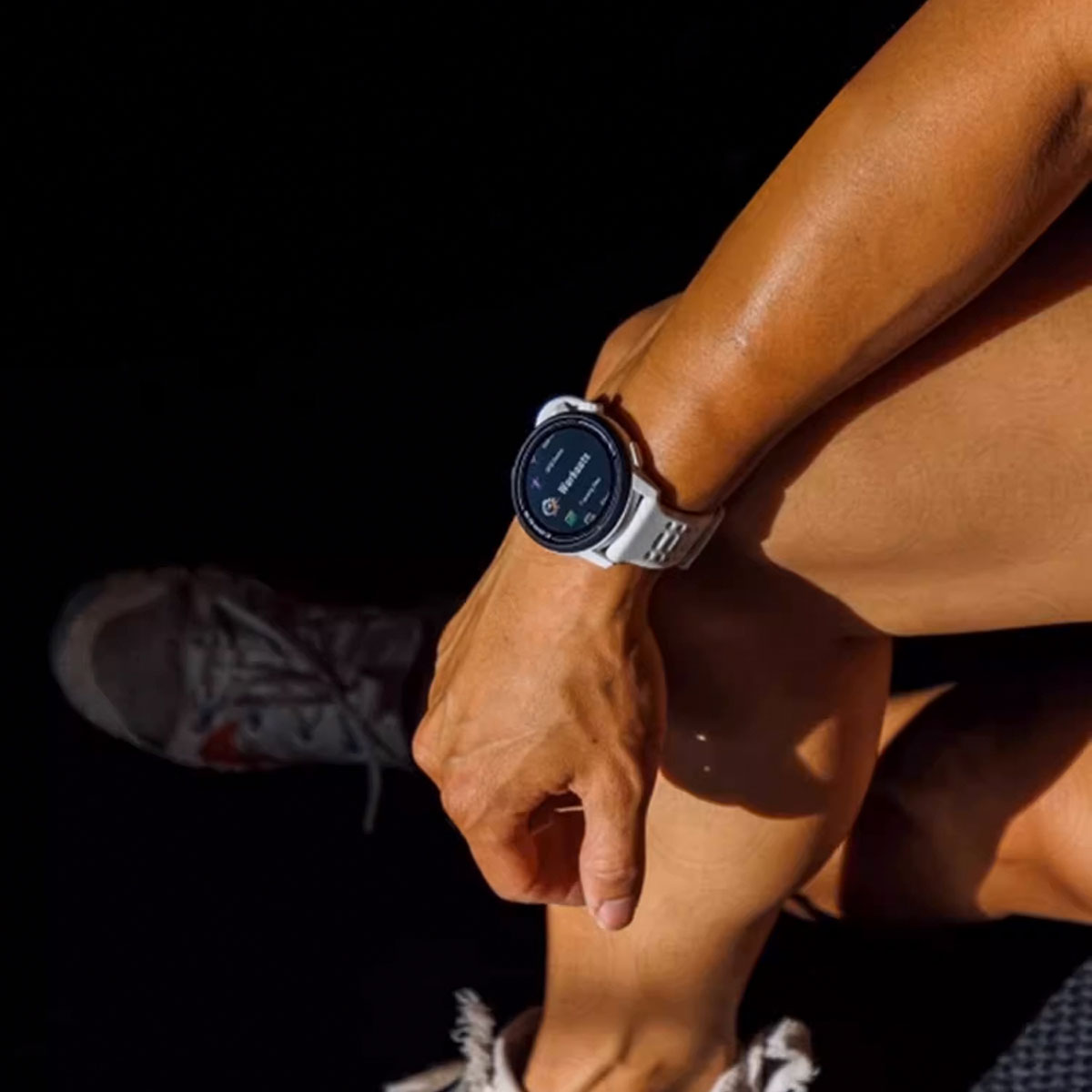 Osoba ma na sobie biały smartwatch. Widoczne jest jej ramię i noga. But do biegania jest w tle. Scena jest ciemna.