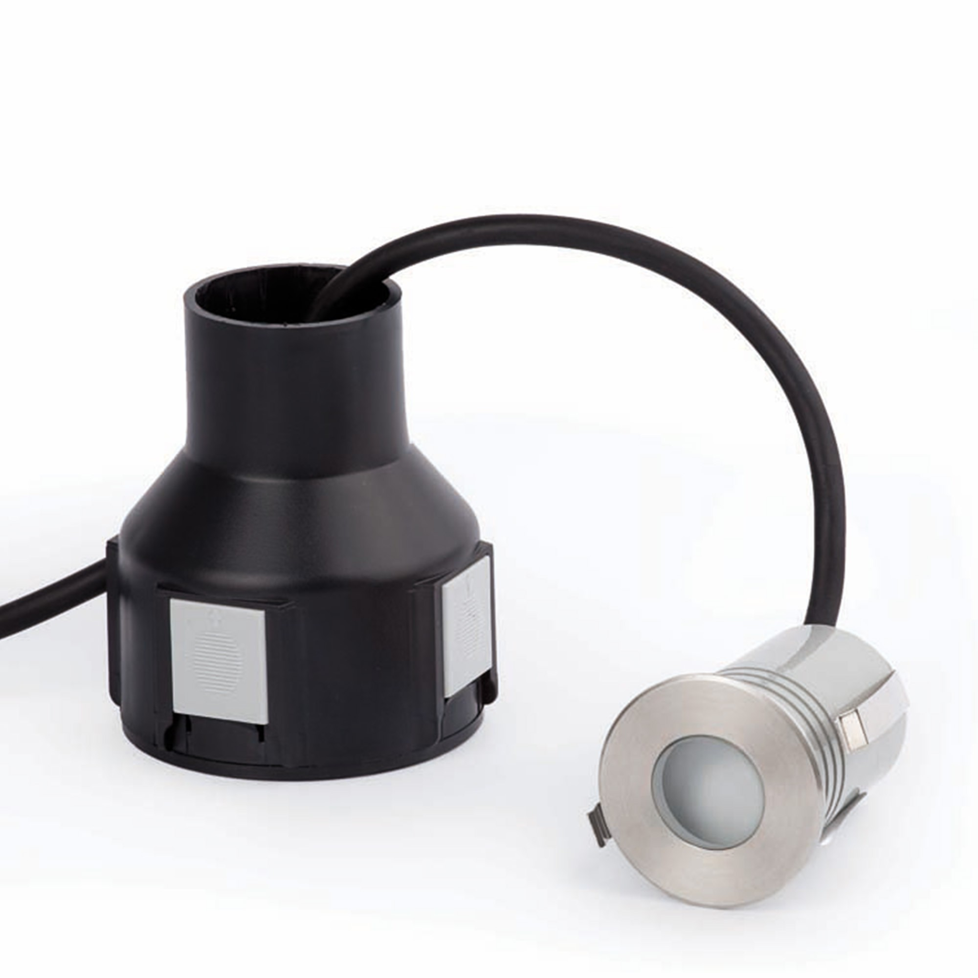 Una base in plastica nera collegata a un apparecchio di illuminazione circolare argentato con un cavo nero curvo. Sfondo bianco.