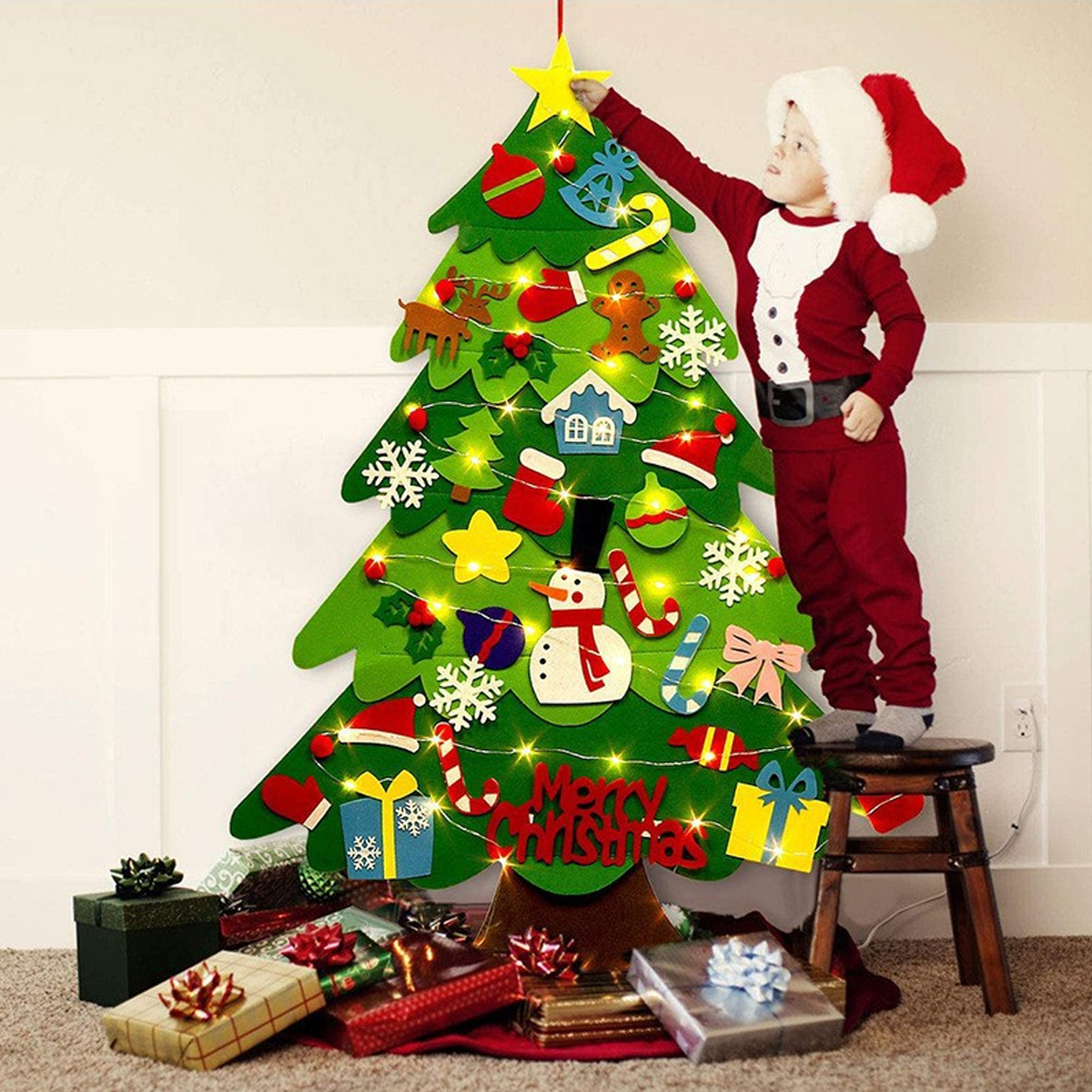 Ein Kind in einem Weihnachtsmannanzug setzt einen Stern auf einen Weihnachtsbaum aus Filz. Geschenke umgeben den Baum auf einem roten Tuch.