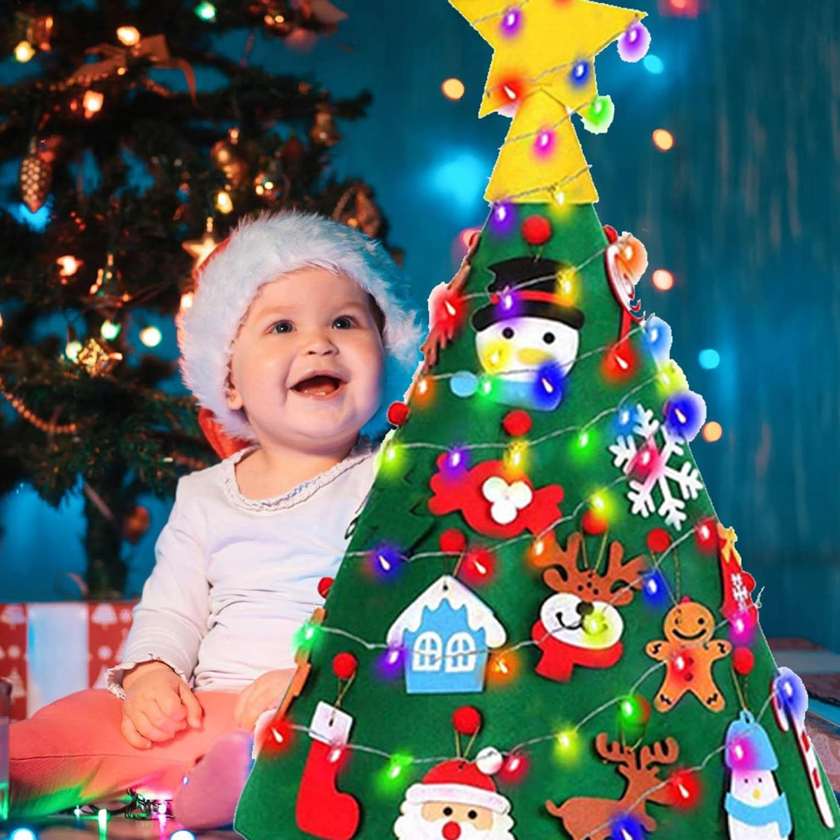 Ein Baby mit Weihnachtsmütze lächelt neben einem Filz-Weihnachtsbaum mit Ornamenten und Lichtern, und einem Weihnachtsbaum im Hintergrund.
