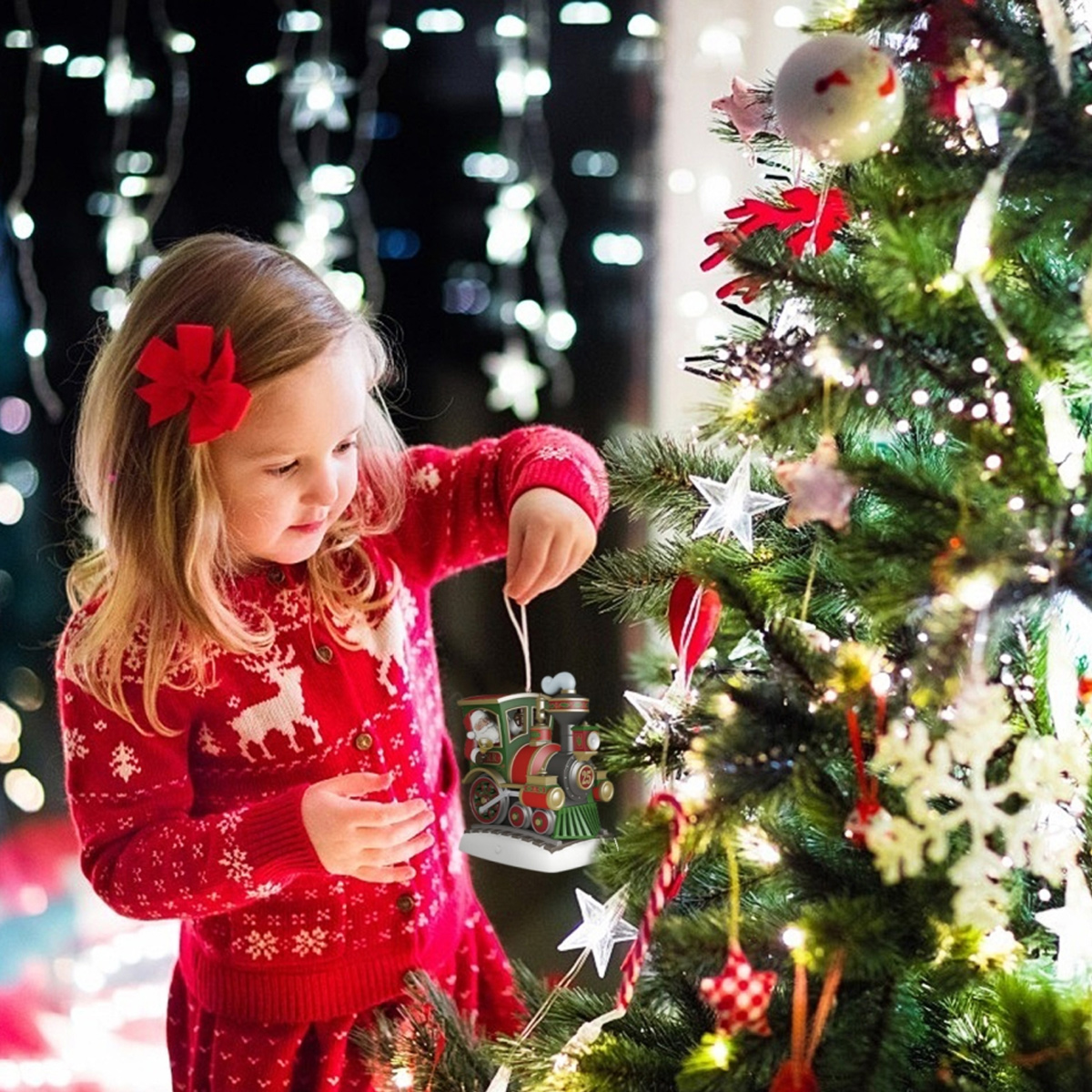 Ein Mädchen in einem roten Pullover schmückt einen Weihnachtsbaum mit einem Zugornament. Der Baum ist mit Lichtern und verschiedenen Ornamenten geschmückt.