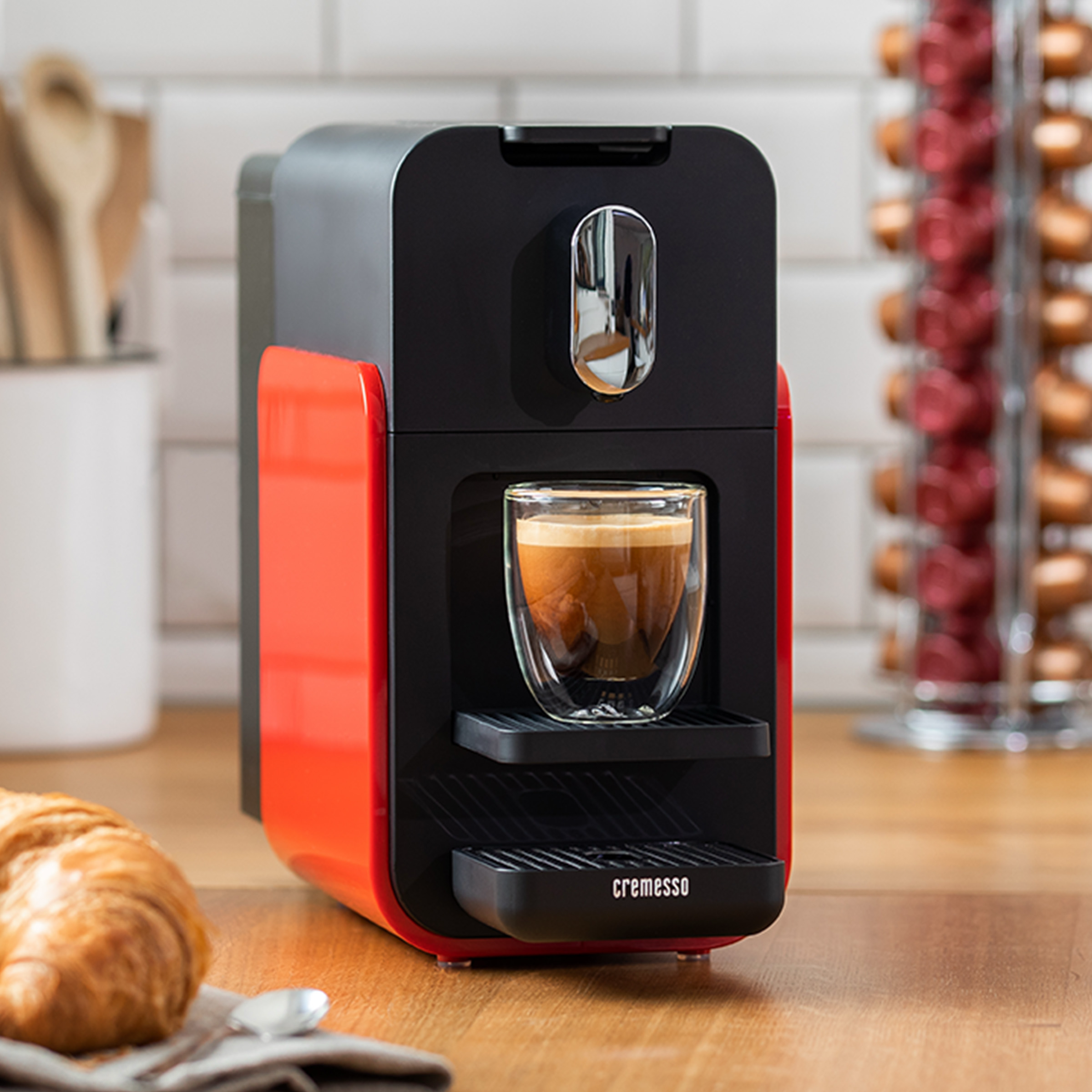 Eine rot-schwarze Kaffeemaschine mit einem Glas Espresso. Ein Croissant und Kaffeepads sind in der Nähe.