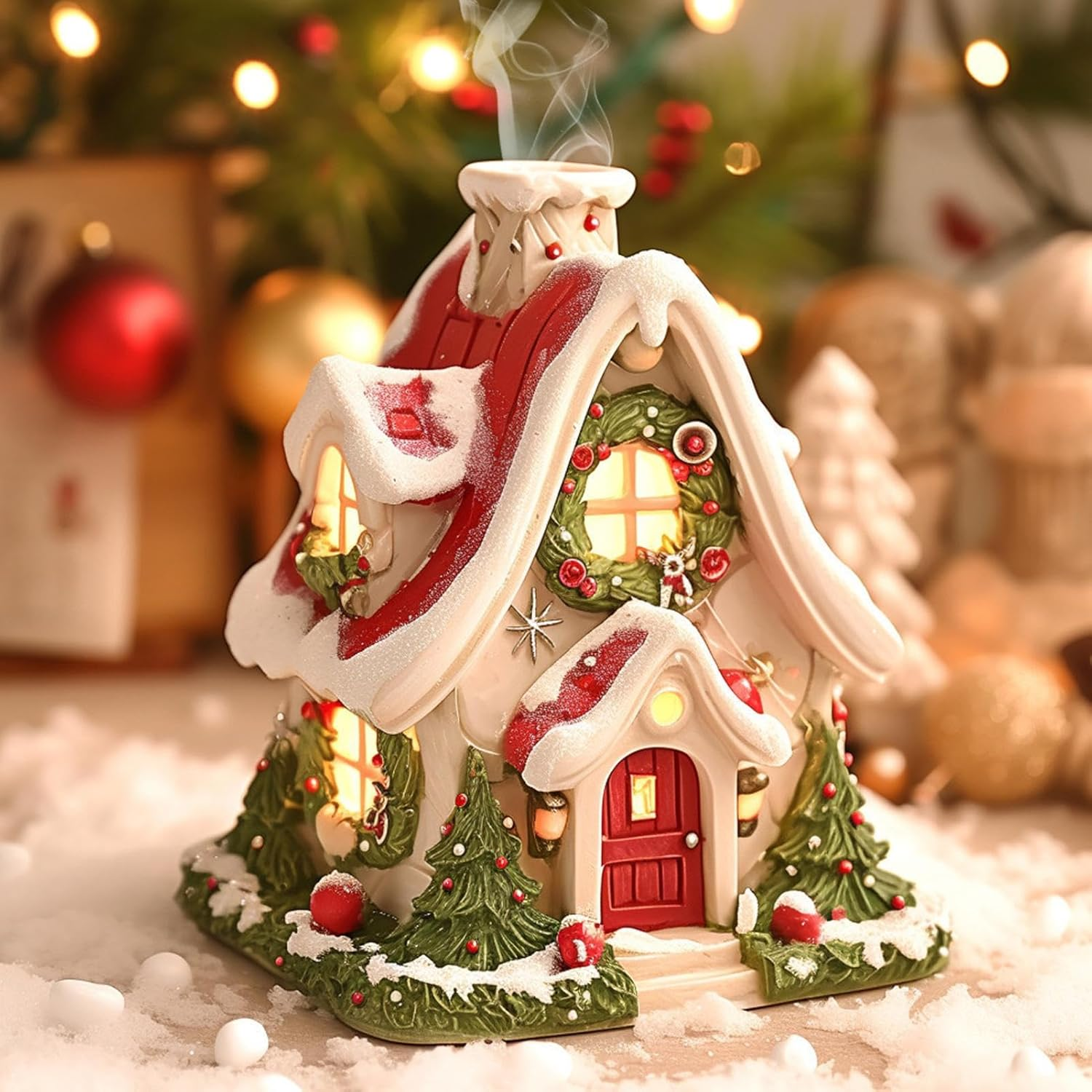 Ein dekoratives Weihnachtshaus. Es hat ein rotes, schneebedecktes Dach, eine rote Tür und ist von Kunstschnee und Ornamenten umgeben.