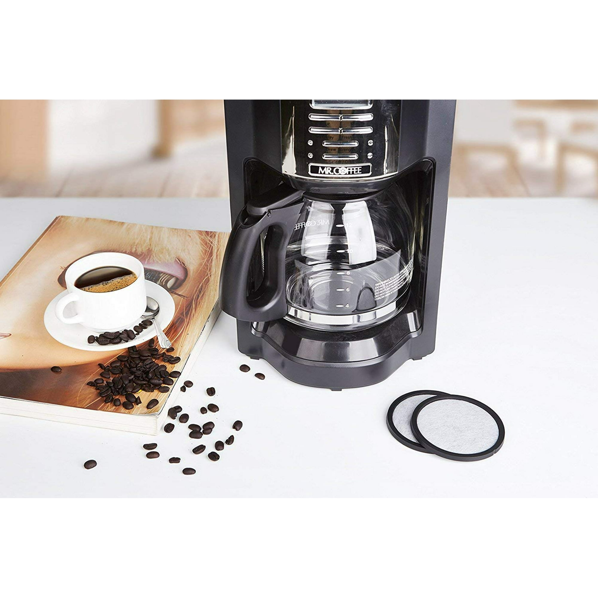 Eine Kaffeemaschine, eine Tasse Kaffee und Kaffeebohnen stehen auf einem Tisch. Filter sind ebenfalls sichtbar.