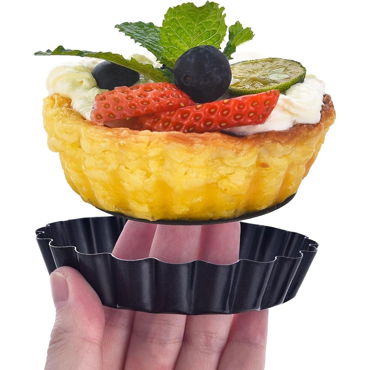 Eine Hand hält eine schwarze Tartpfanne unter einem Obstkuchen mit Erdbeeren, Blaubeeren, Sahne und Minze.