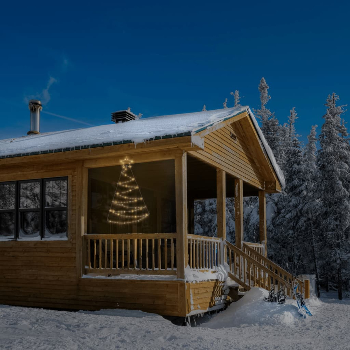 Holzhütte in verschneiter Landschaft mit beleuchtetem Weihnachtsbaumumriss im Fenster. Schneebedecktes Dach und Bäume. Klarer blauer Himmel.