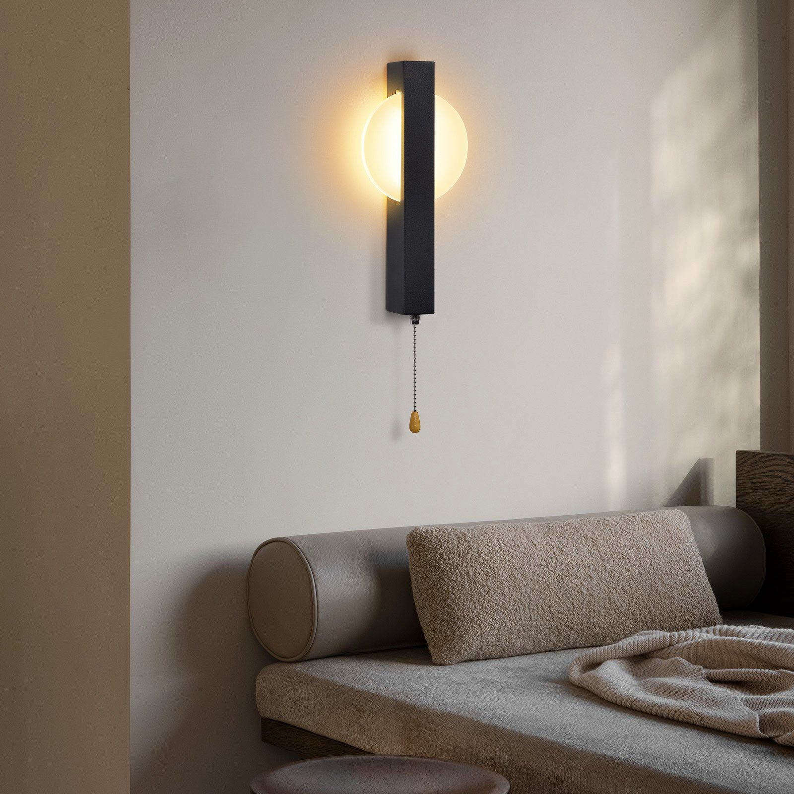 Una moderna lampada da parete con catena pende sopra un divano con cuscini e una coperta. La lampada ha una luce rotonda.