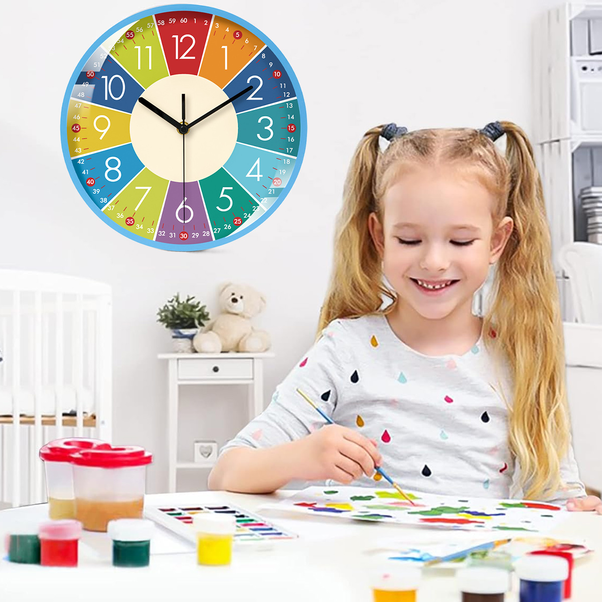 Ein Mädchen malt am Tisch. Eine bunte Uhr hängt an der Wand. Farben und Pinsel liegen auf dem Tisch. Ein Teddybär sitzt daneben.