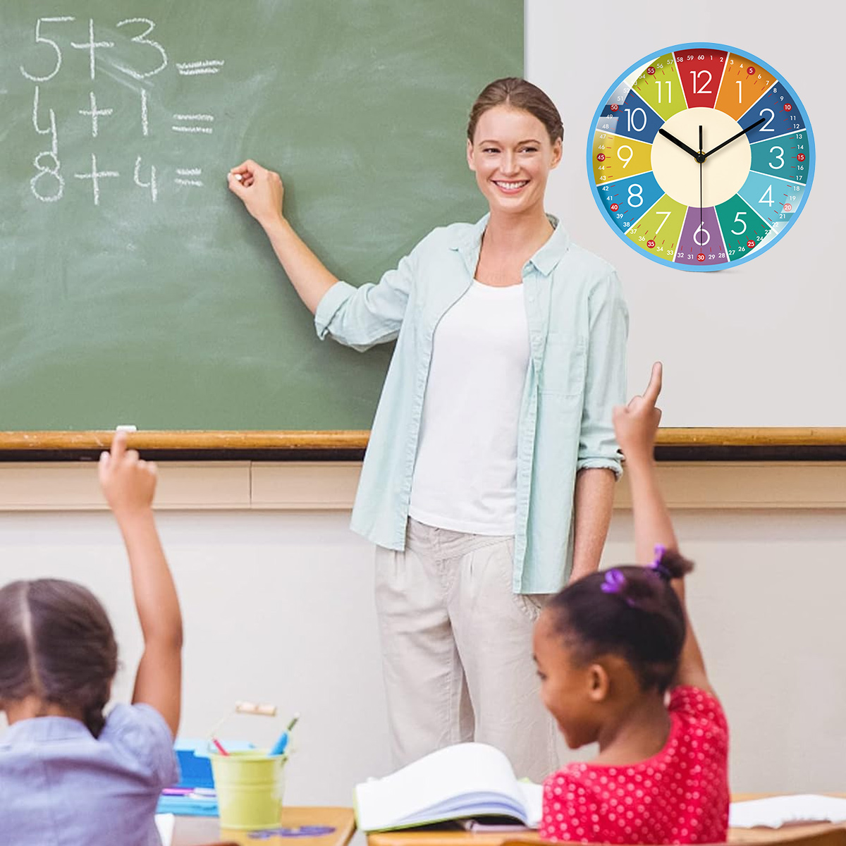 Eine Lehrerin zeigt auf eine Tafel mit Matheaufgaben. Schüler heben die Hände. Eine bunte Uhr hängt an der Wand.