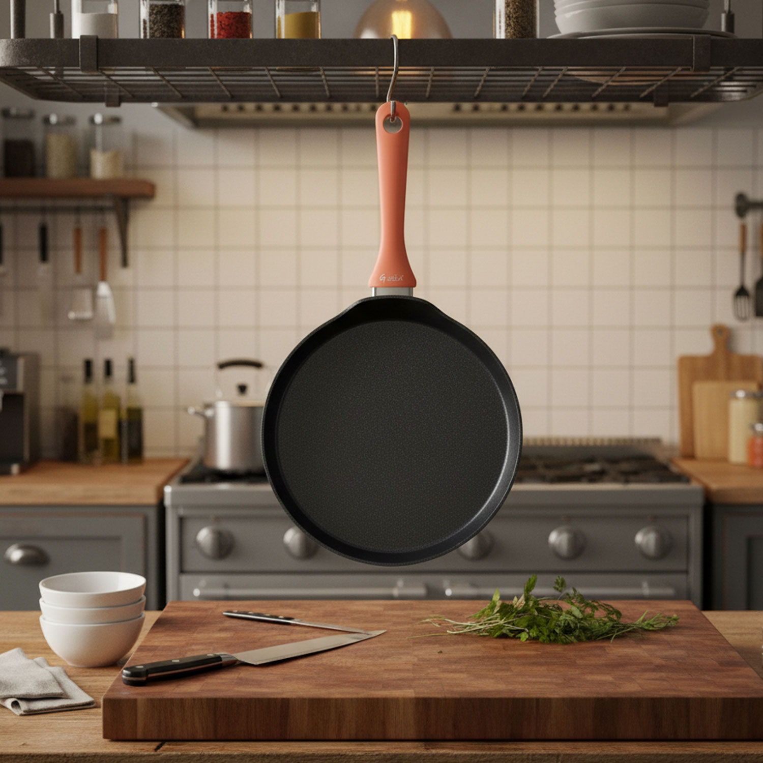 Una sartén redonda negra con mango melocotón cuelga en una cocina. Una tabla de cortar de madera con cuchillo y verduras está en una encimera.