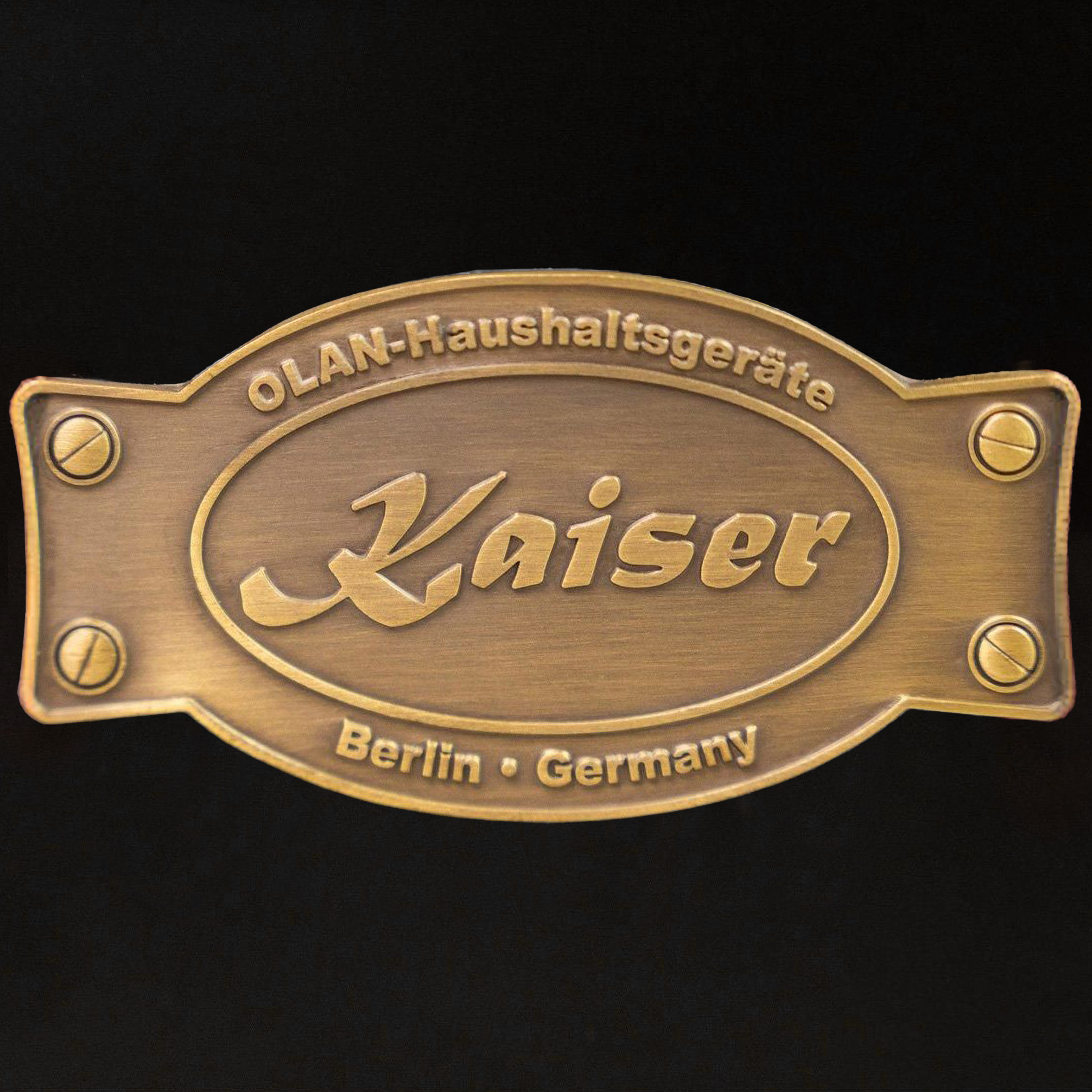 Eine bronzene Kaiser-Plakette mit Text: OLAN-Haushaltgeräte, Kaiser, Berlin, Deutschland. Schrauben befestigen die Plakette auf schwarzem Grund.
