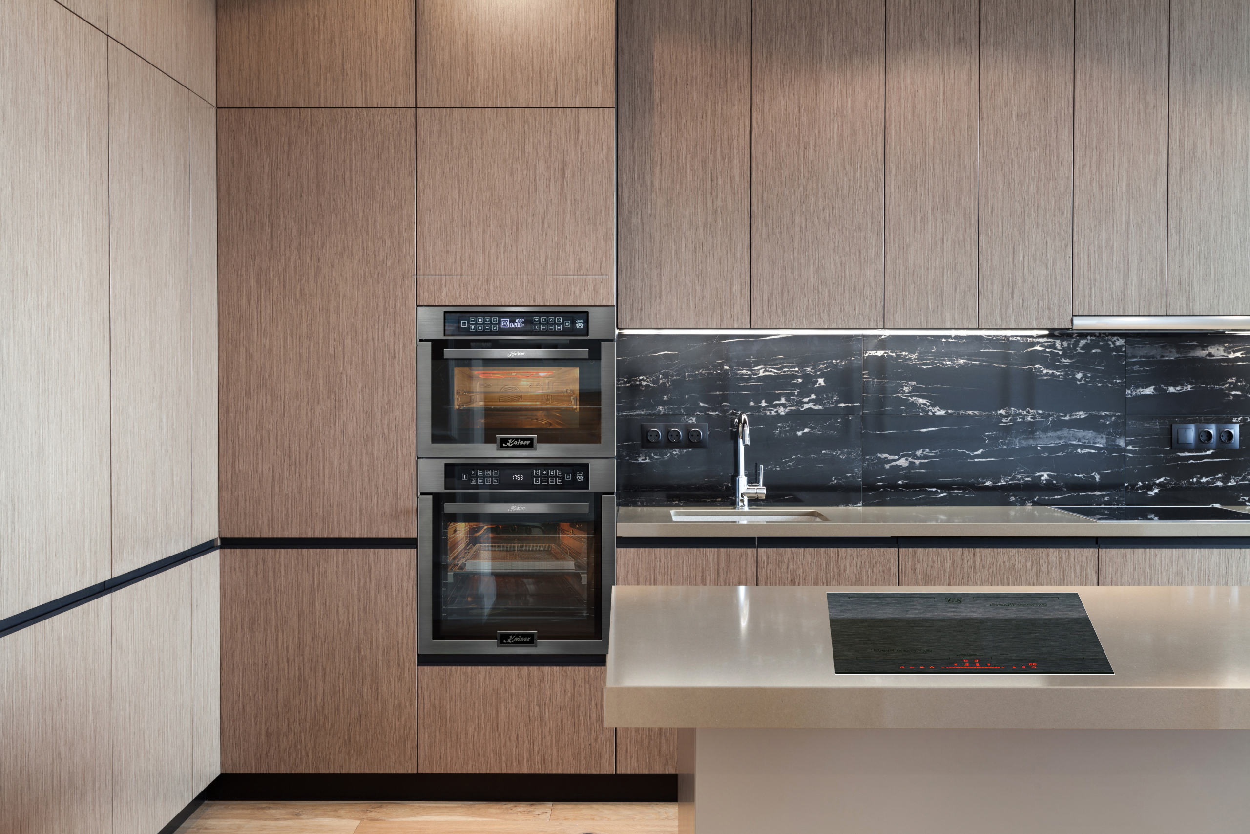 Cucina moderna con forni, piano di lavoro e alzatina in marmo nero. Sono visibili anche mobili in legno e un piano cottura.