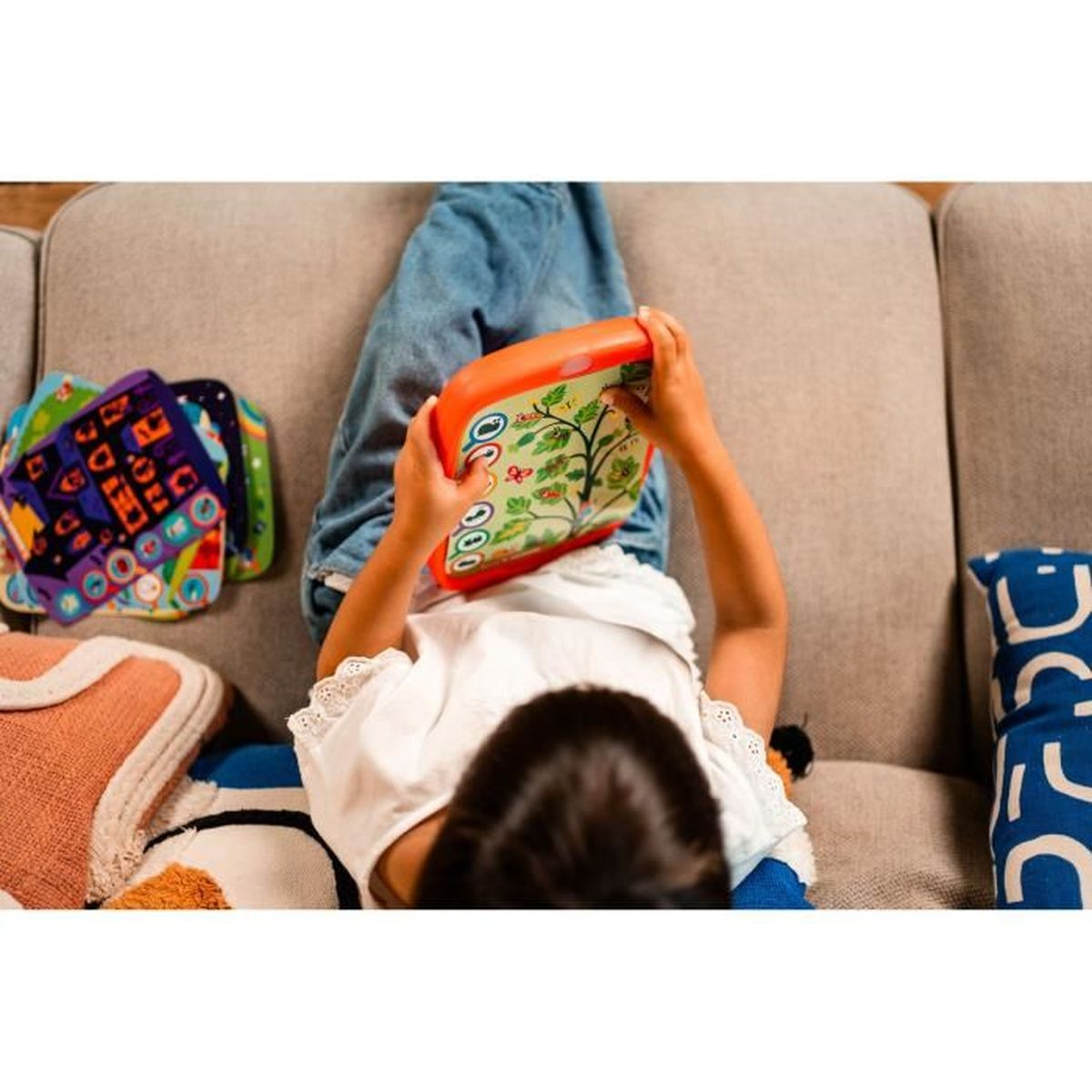 Ein Kind liegt auf einem Sofa und hält ein orangefarbenes Tablet mit einem Baumdesign. Bunte Kissen und blaue Jeans sind ebenfalls zu sehen.