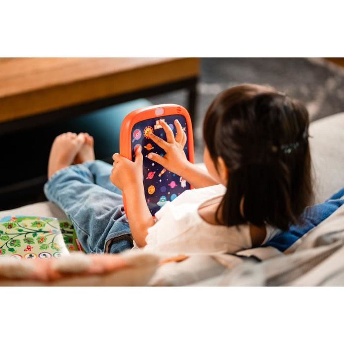 Ein Kind liegt und spielt mit einem Tablet. Das Tablet zeigt eine Weltraumszene. Das Kind trägt Jeans und ein weißes Hemd.