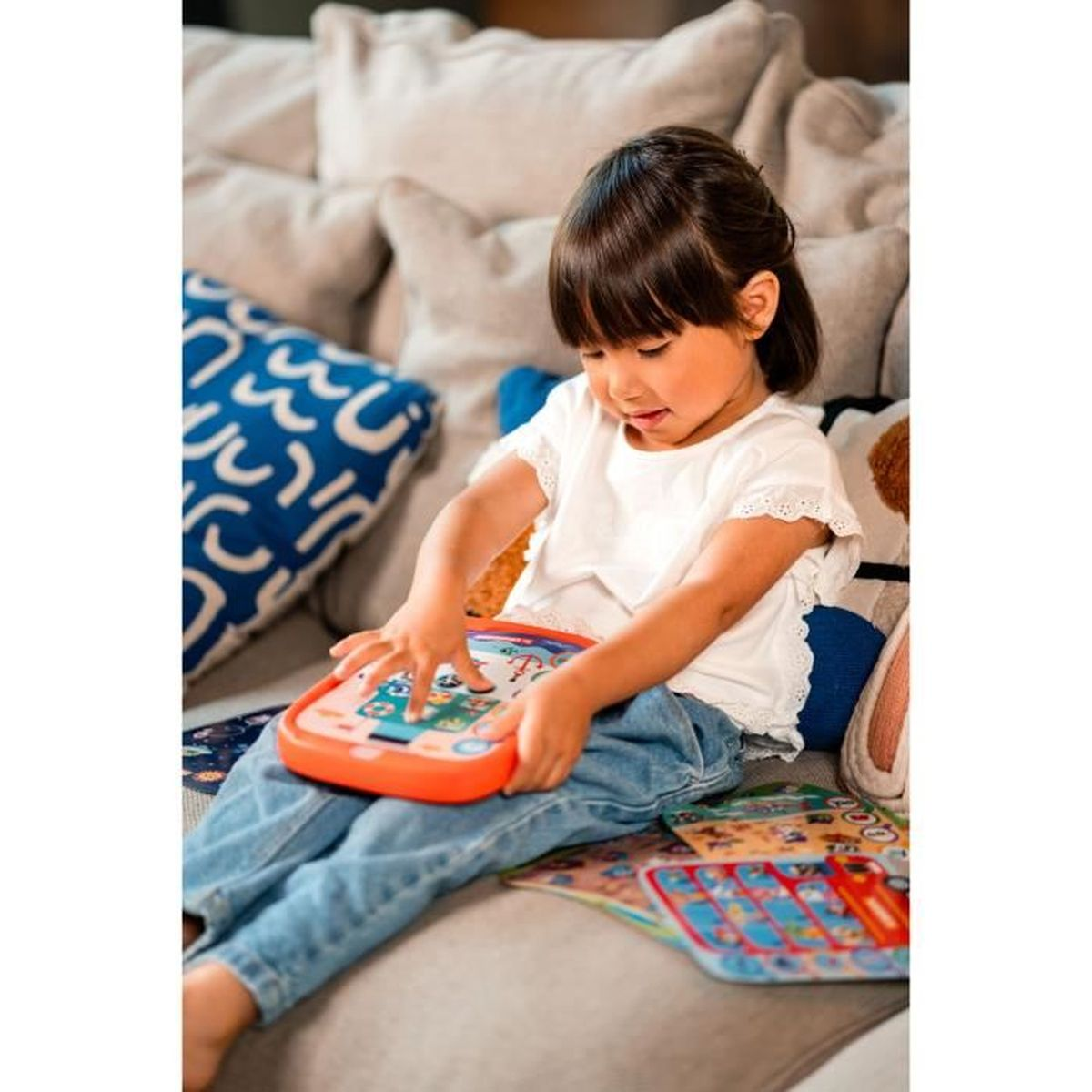 Ein junges Mädchen sitzt auf einer Couch und spielt mit einem Spielzeugtablett. Sie trägt ein weißes Hemd und Jeans. Kissen sind im Hintergrund.