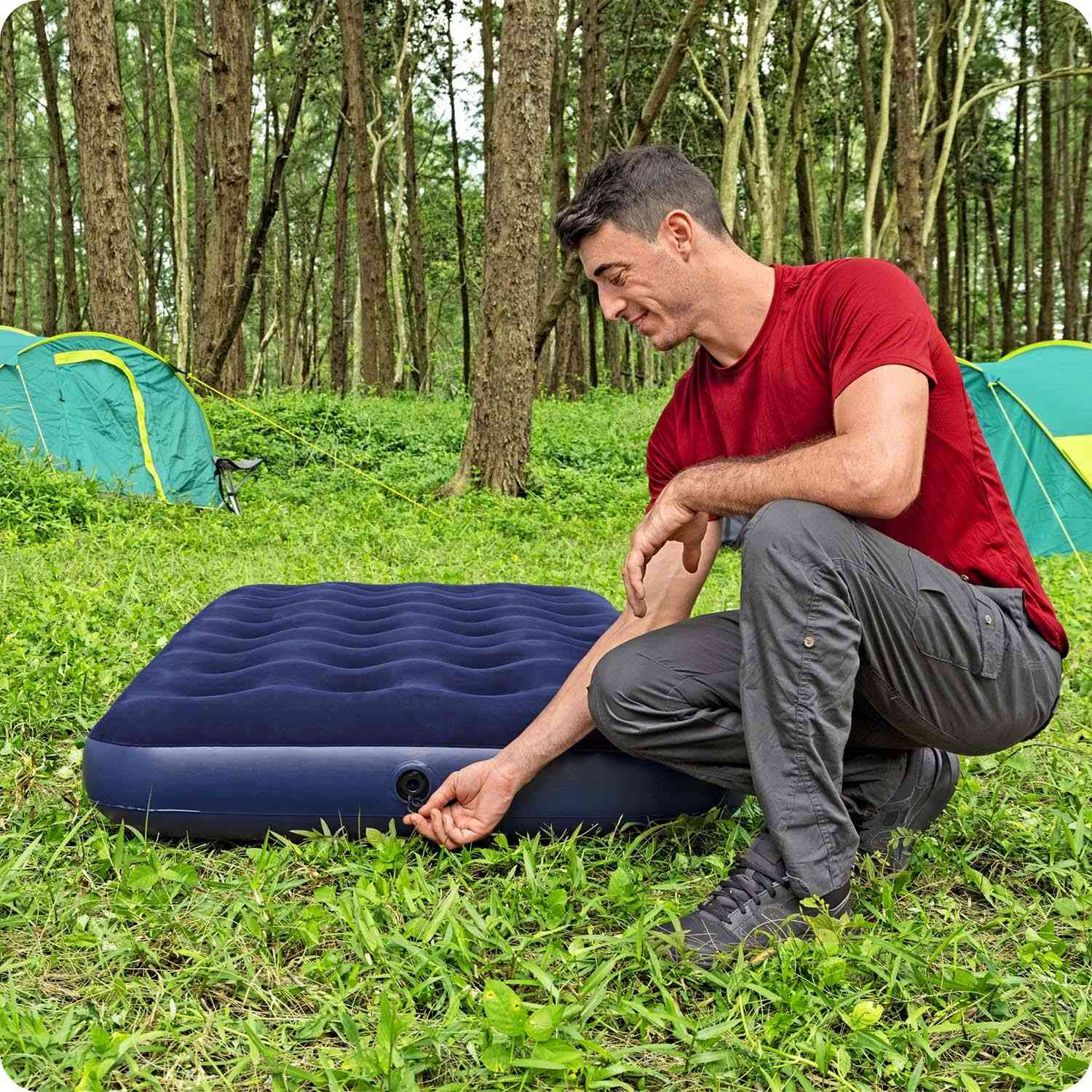 Un hombre con una camisa roja se arrodilla junto a un colchón inflable azul en un bosque. Está ajustando una válvula. Hay tiendas verdes al fondo.