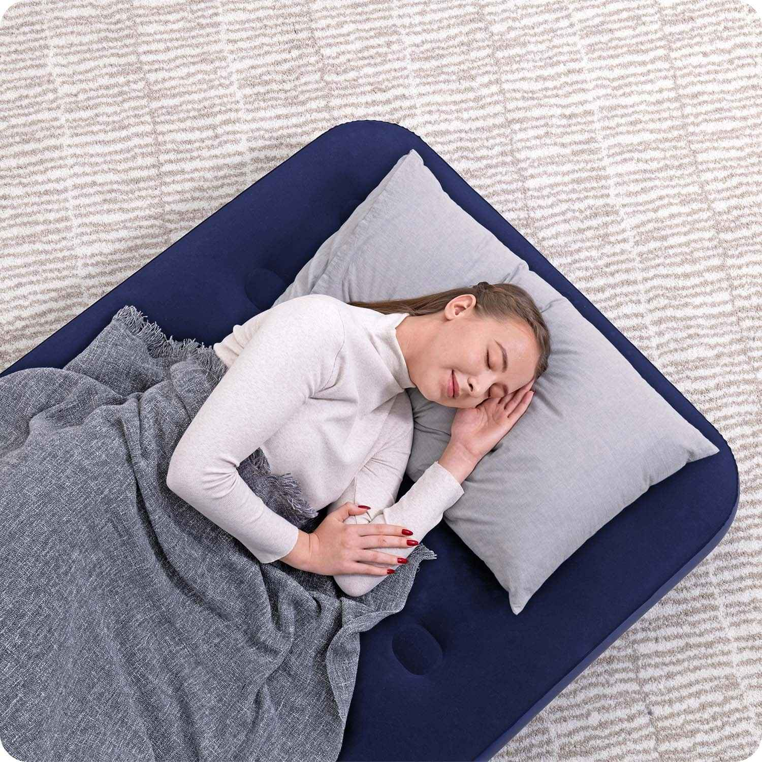 Una mujer duerme en un colchón de aire azul marino con una almohada y una manta grises. El fondo es una alfombra estampada.